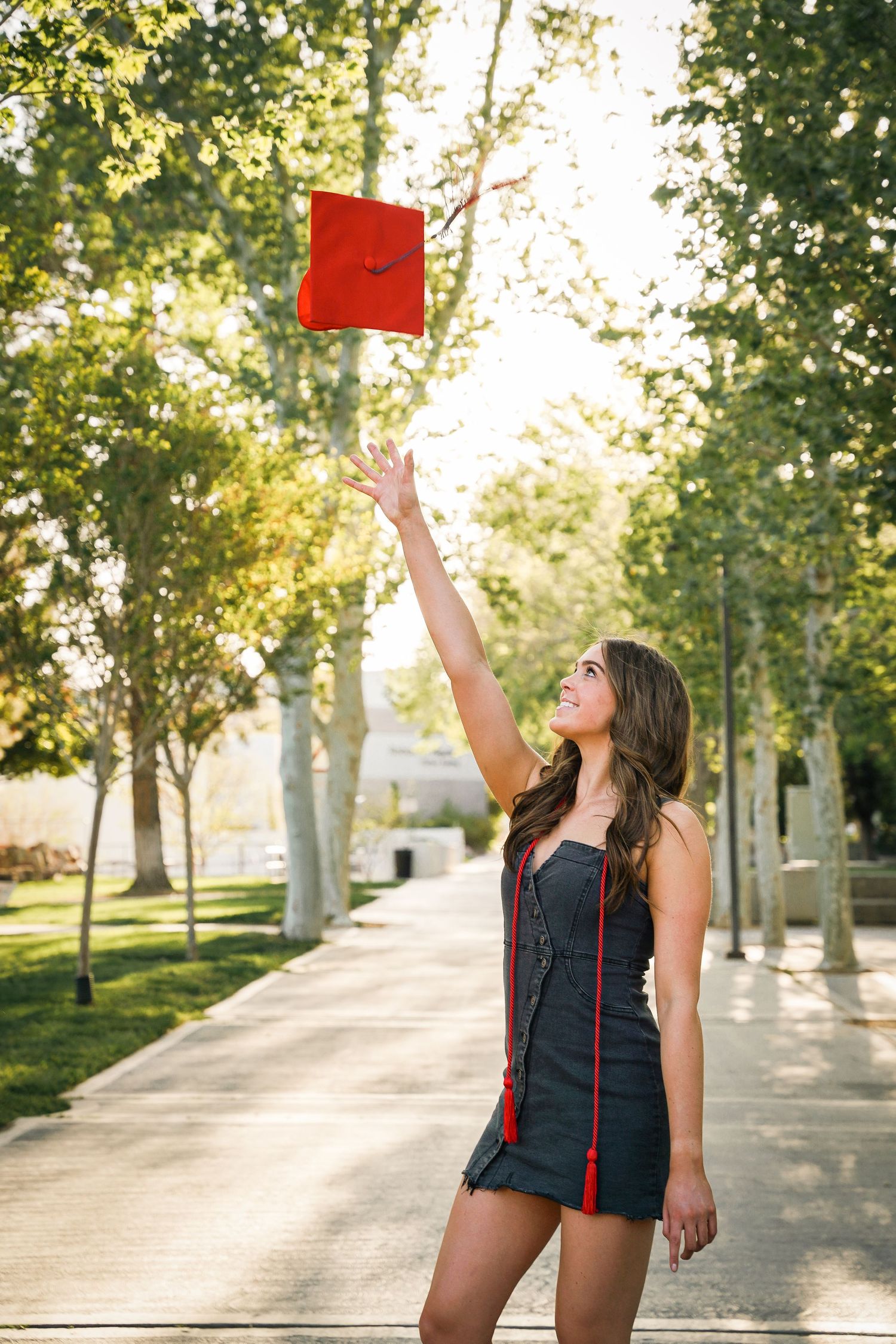 Graduate joyfully tosses red diploma holder in air on sunny tree-lined campus path.