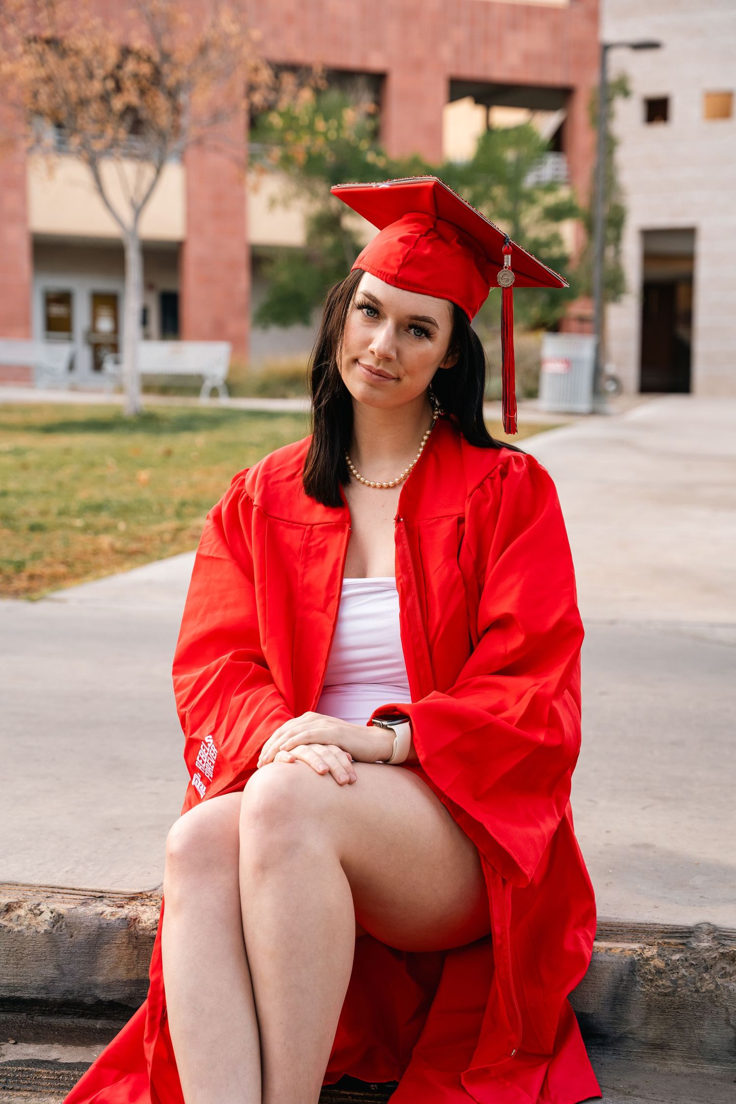 Graduate in red cap and gown sitting on stone wall outdoors.