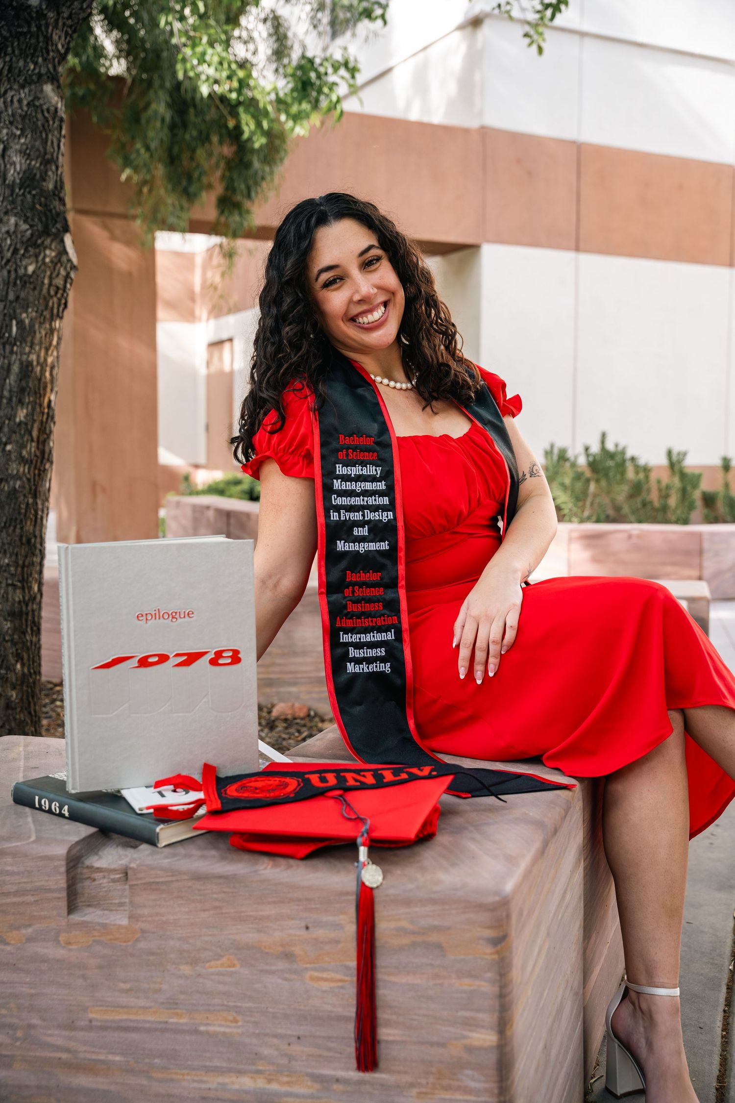 Graduate posing in a vibrant red dress with a graduation stole on a sunny outdoor campus.