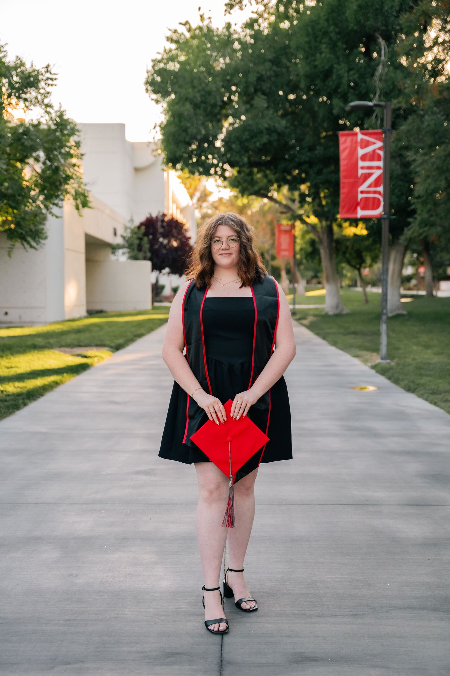 Graduate in black dress carries red diploma folder while walking on UNLV campus pathway.