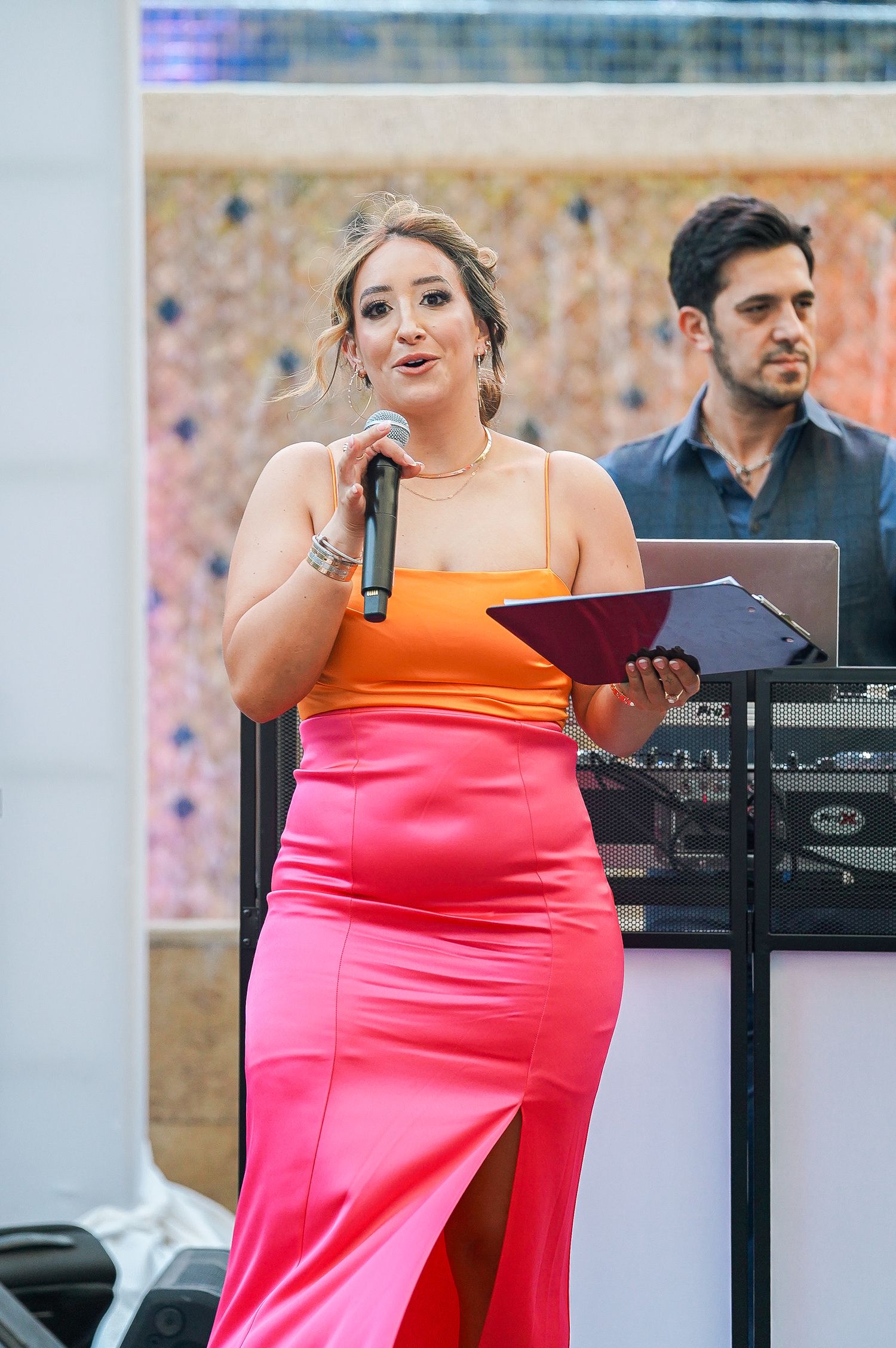 A performer in orange and pink dress sings with a microphone at an event.