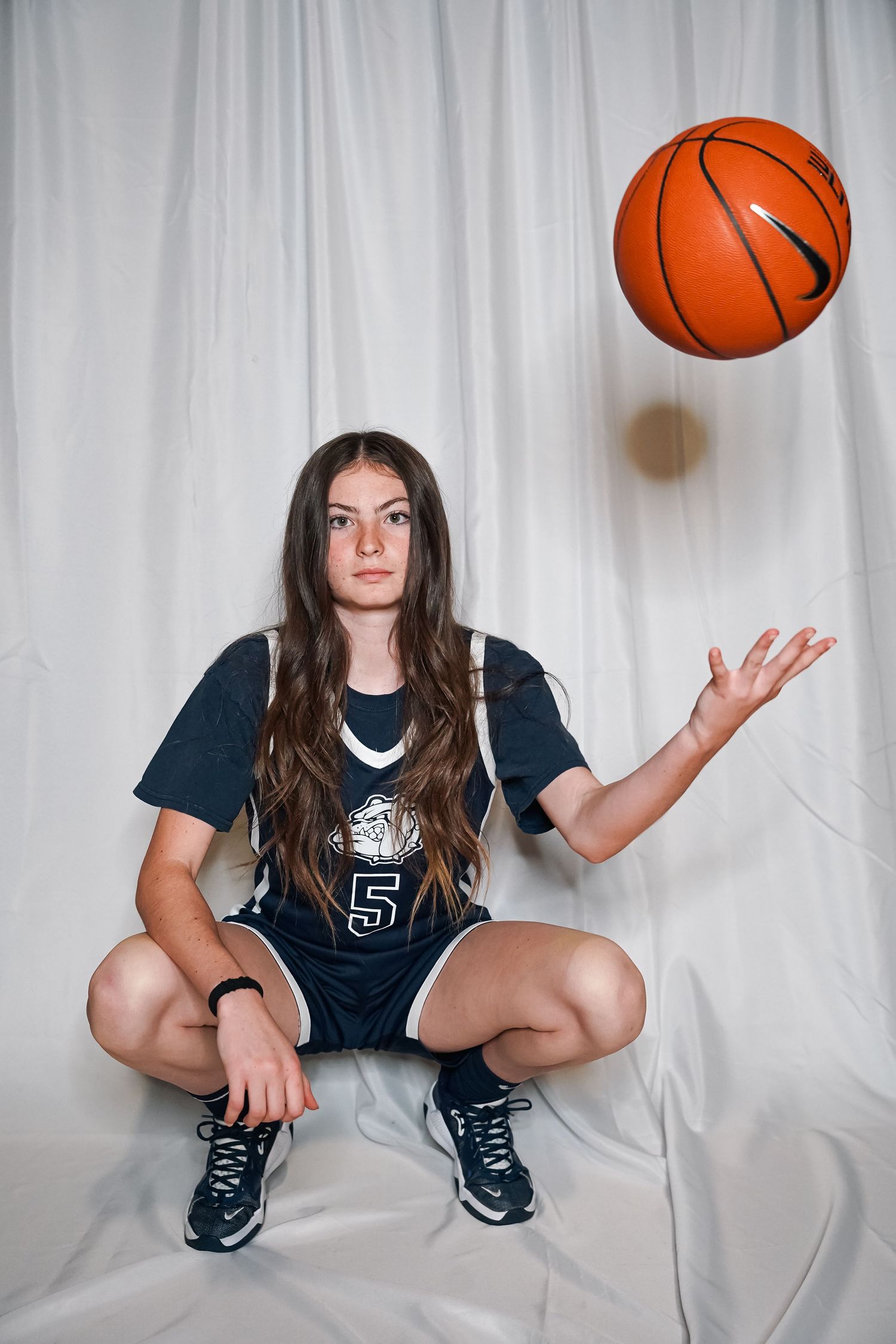 Basketball player in navy uniform tosses ball while crouching against white backdrop.