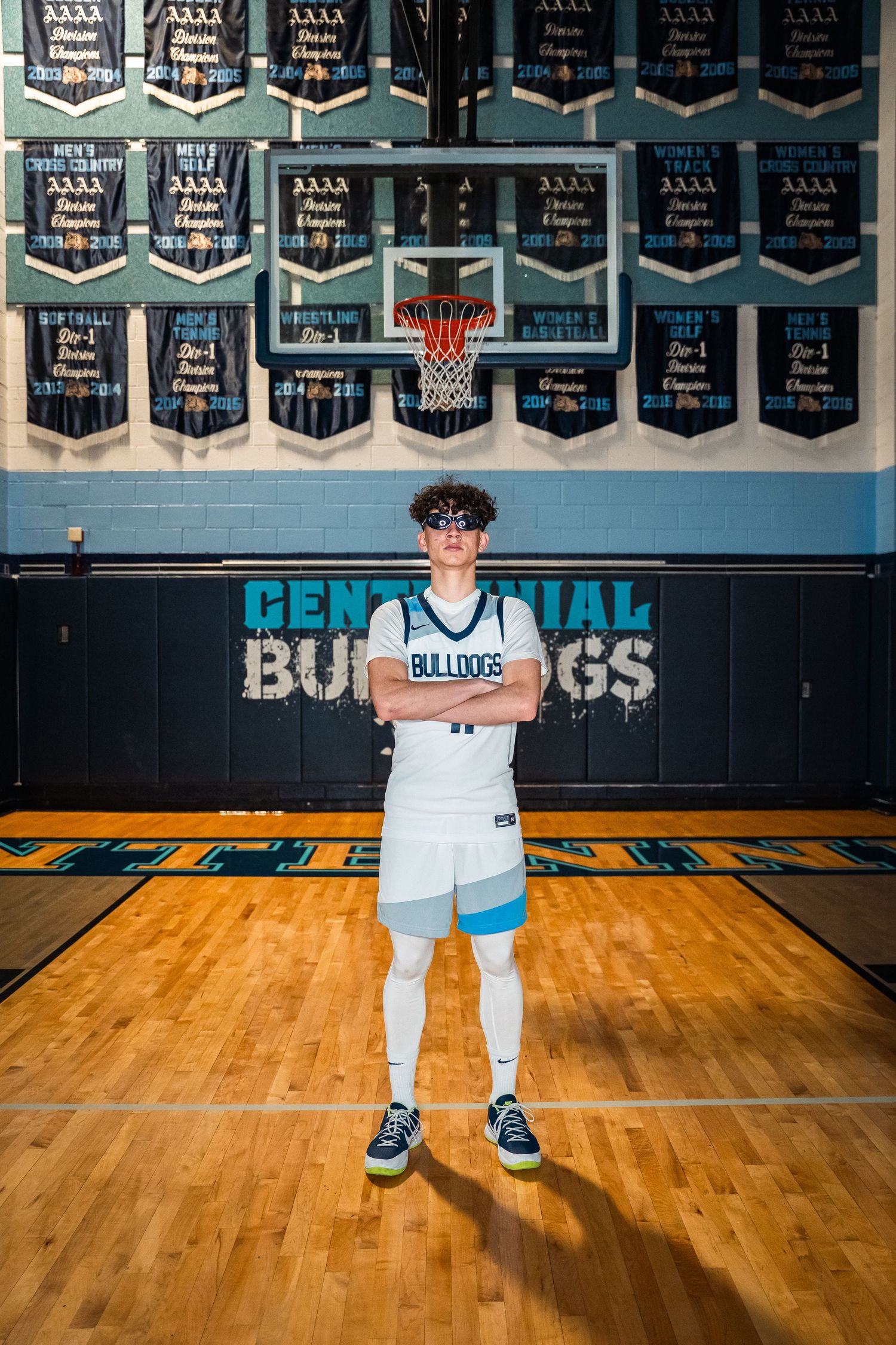 Basketball player in white Bulldogs uniform stands with arms crossed on gymnasium court.