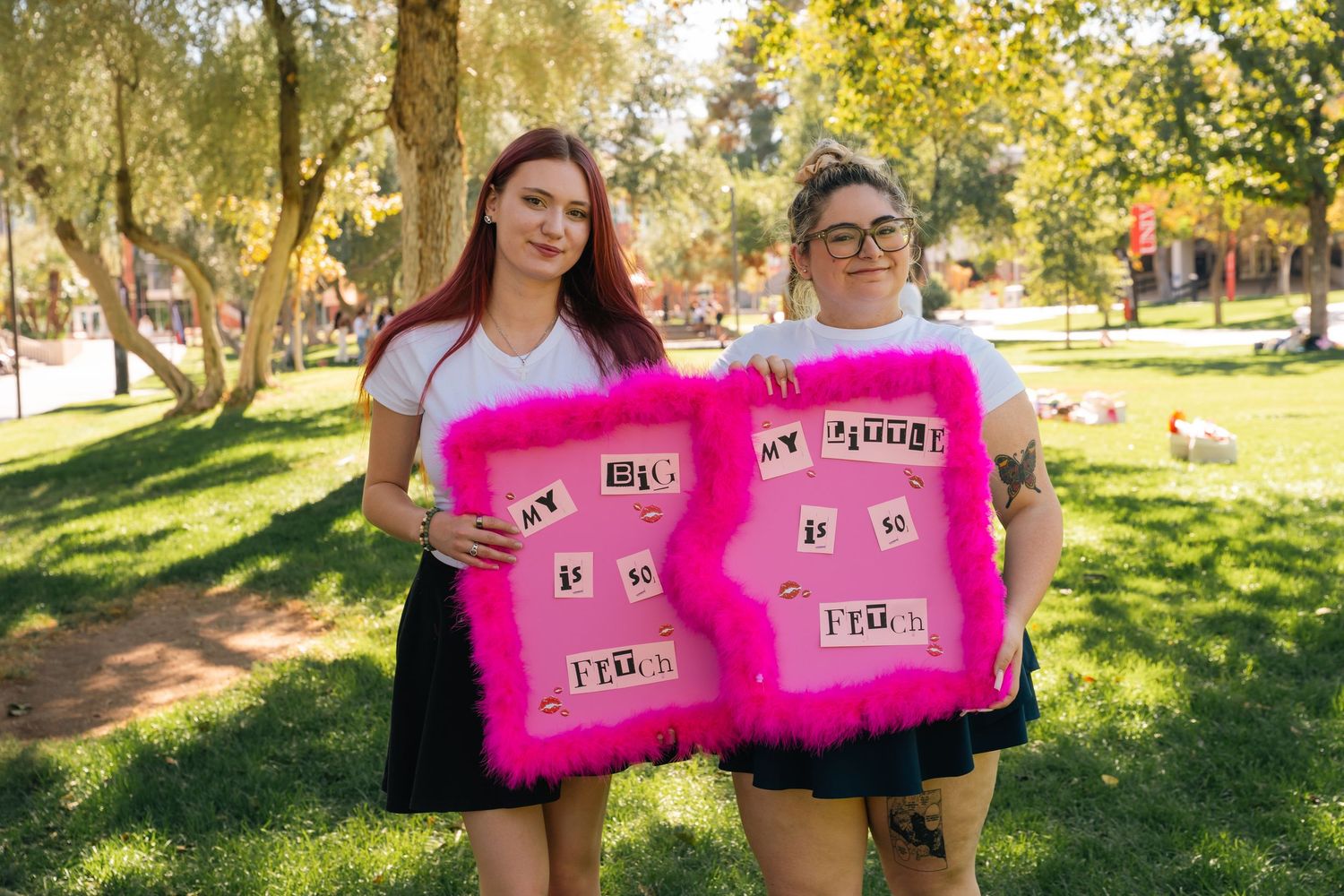 Two graduates hold pink decorated signs on sunny campus lawn.