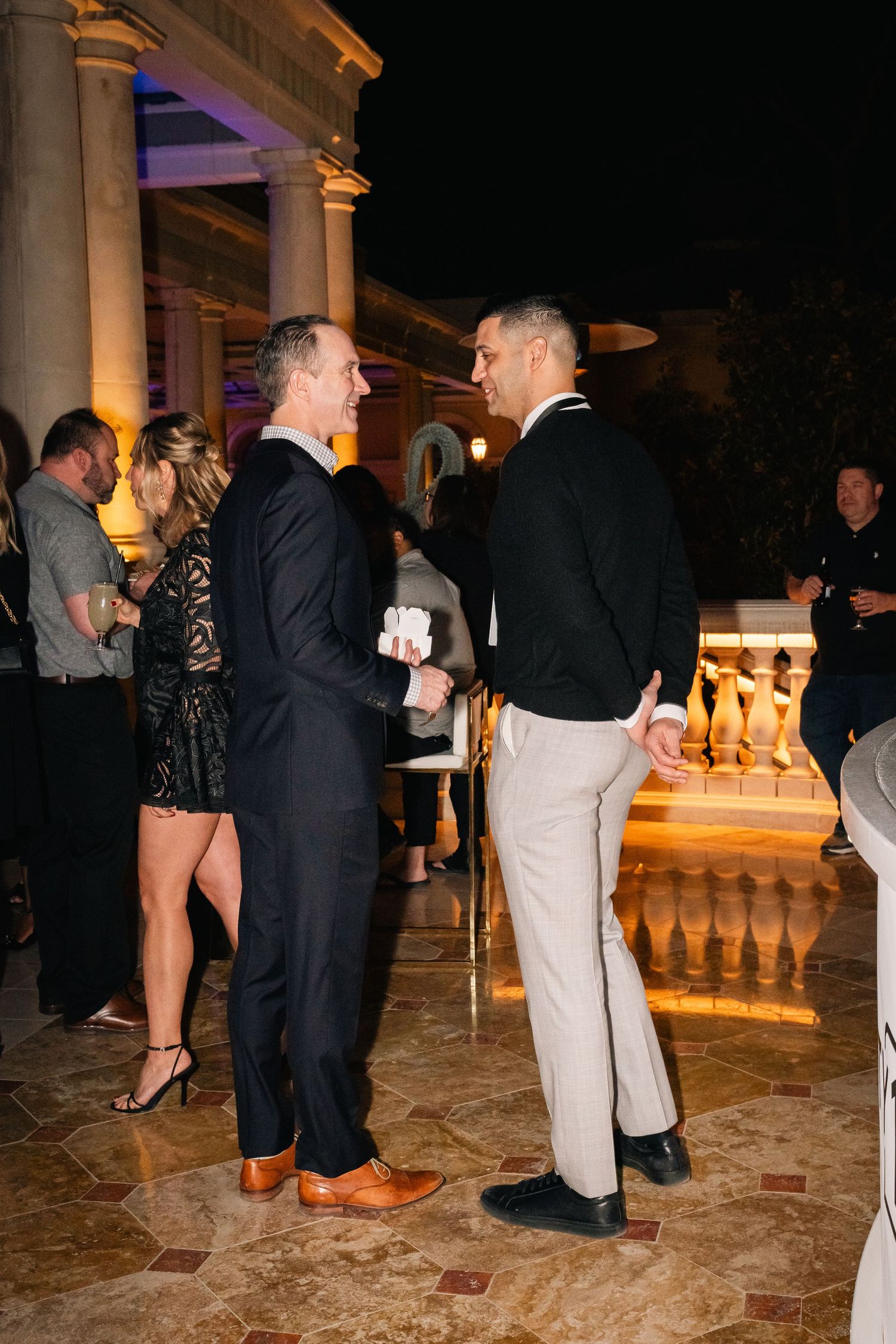 Formal event attendees engage in conversation on a stone patio near classical columns.