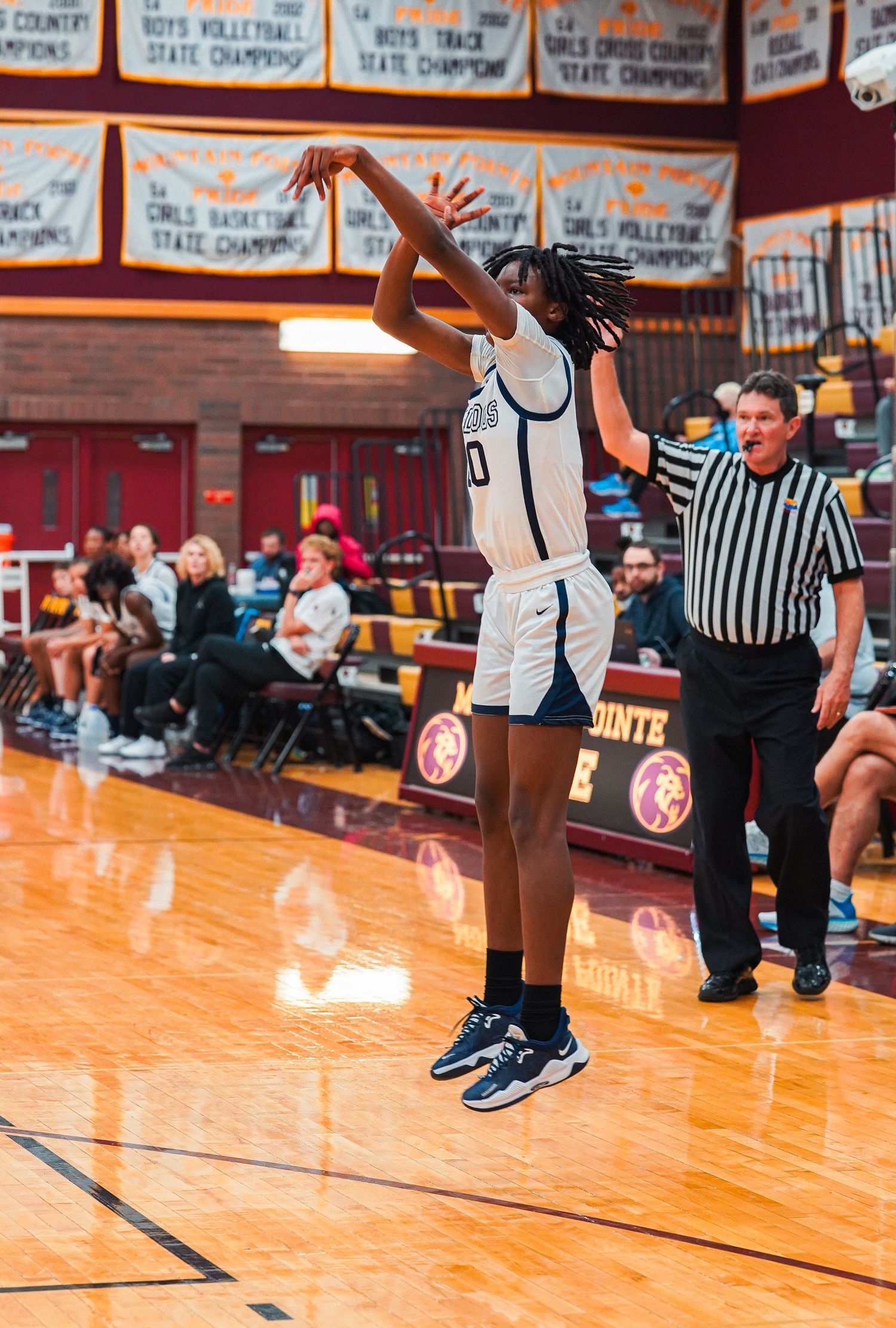 Basketball player takes jump shot while referee watches in gymnasium with championship banners.