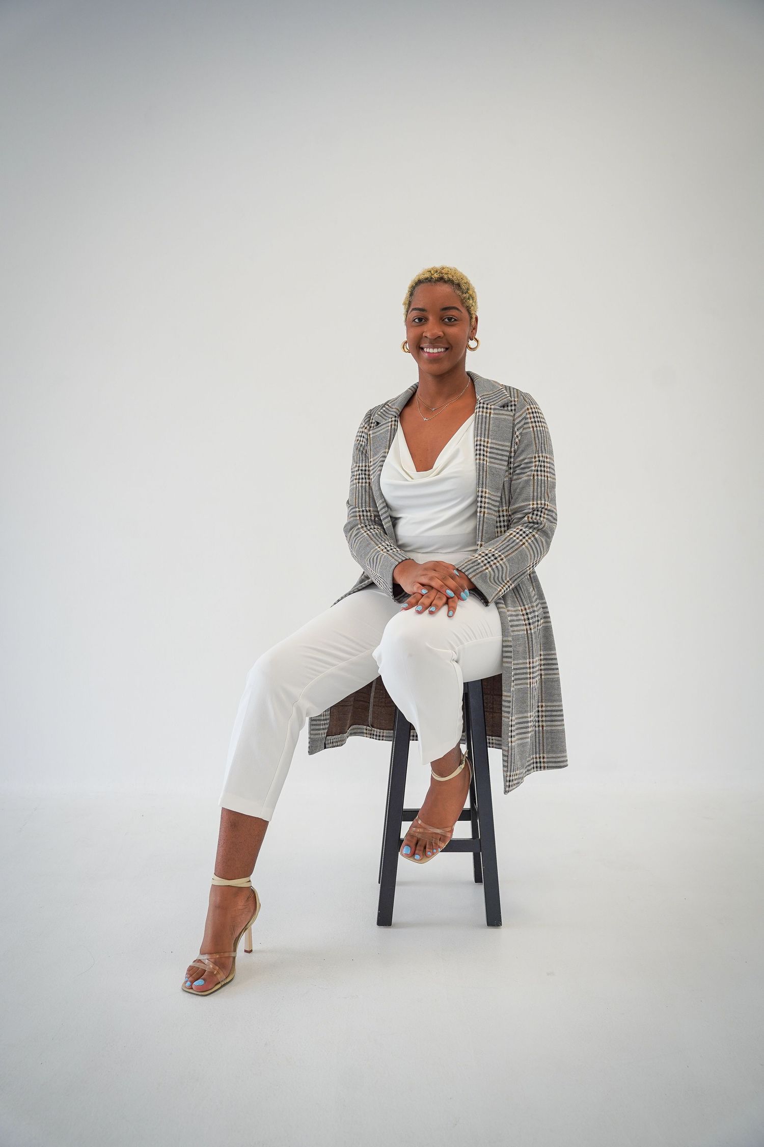 Monaco Media. Professional studio portrait of a woman in white attire and plaid coat sitting on a black stool.