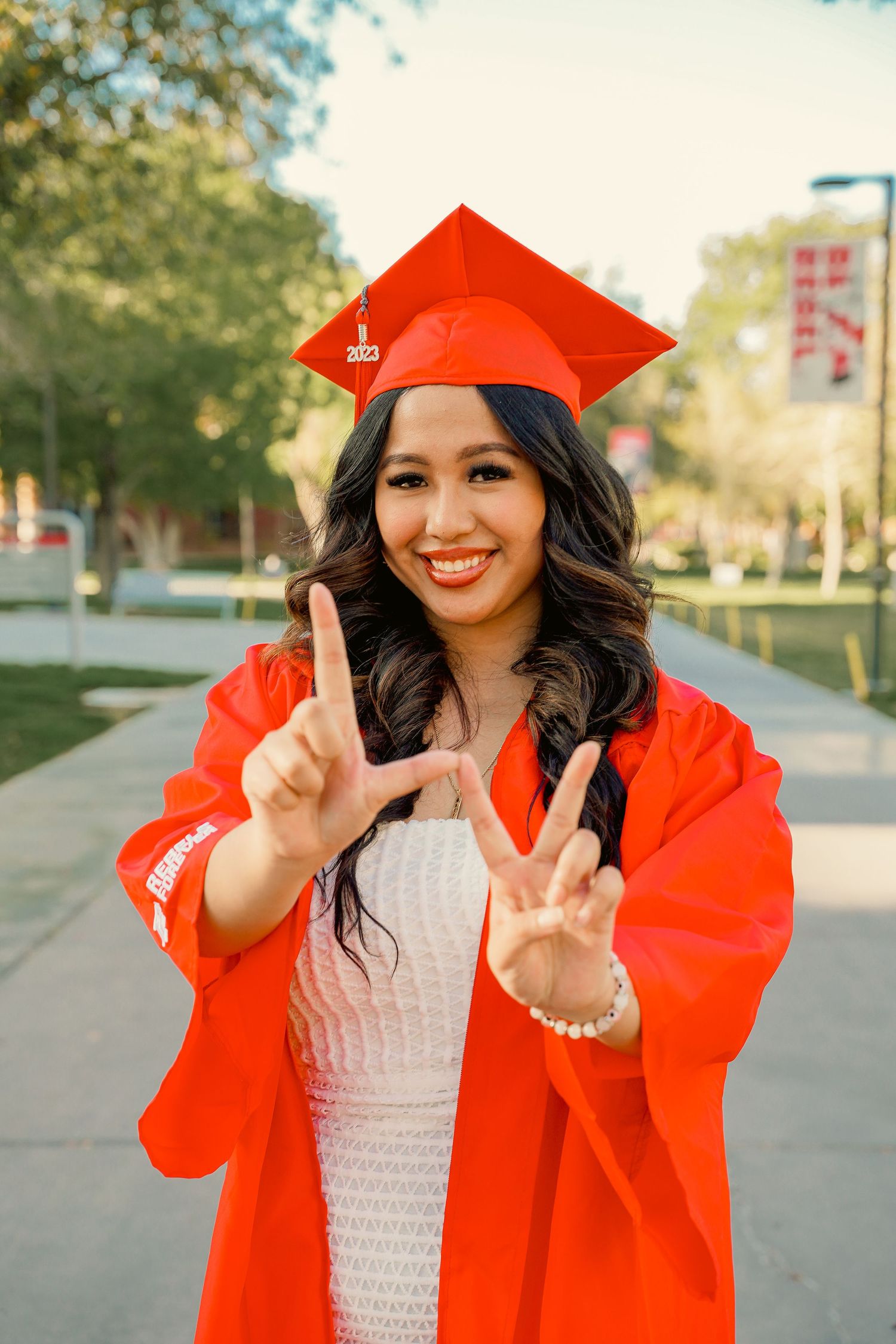 Graduate in bright red cap and gown making peace signs while celebrating on campus pathway.
