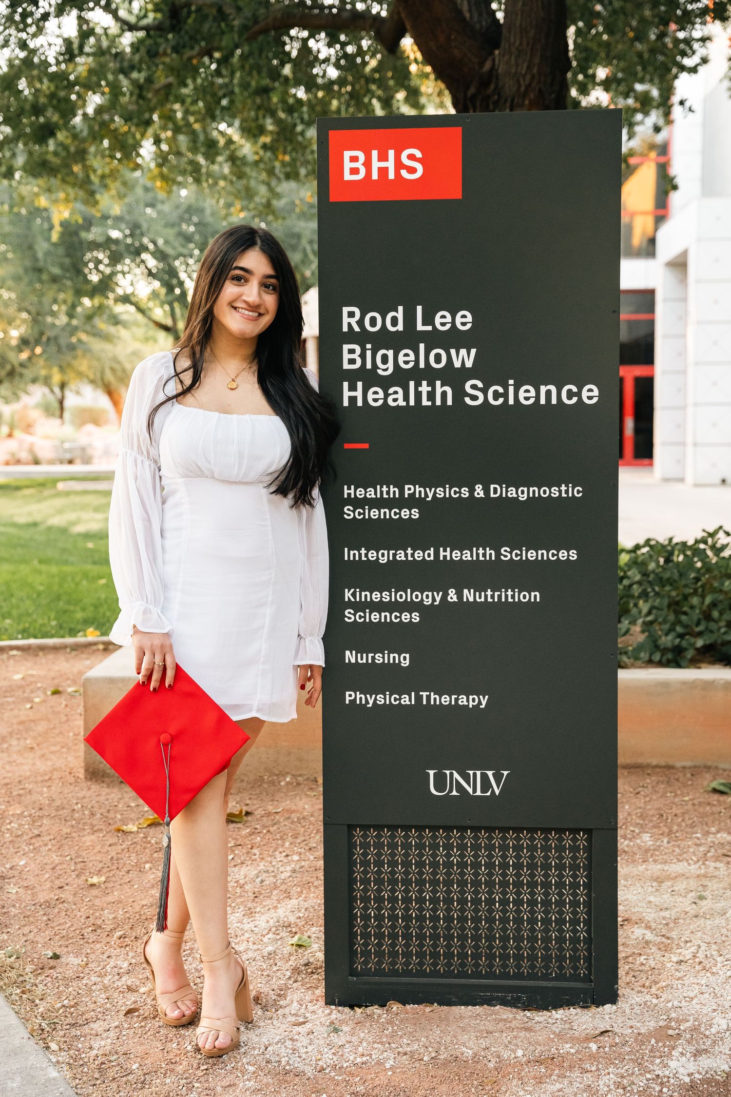 Graduate in white dress posing next to UNLV health sciences building sign.