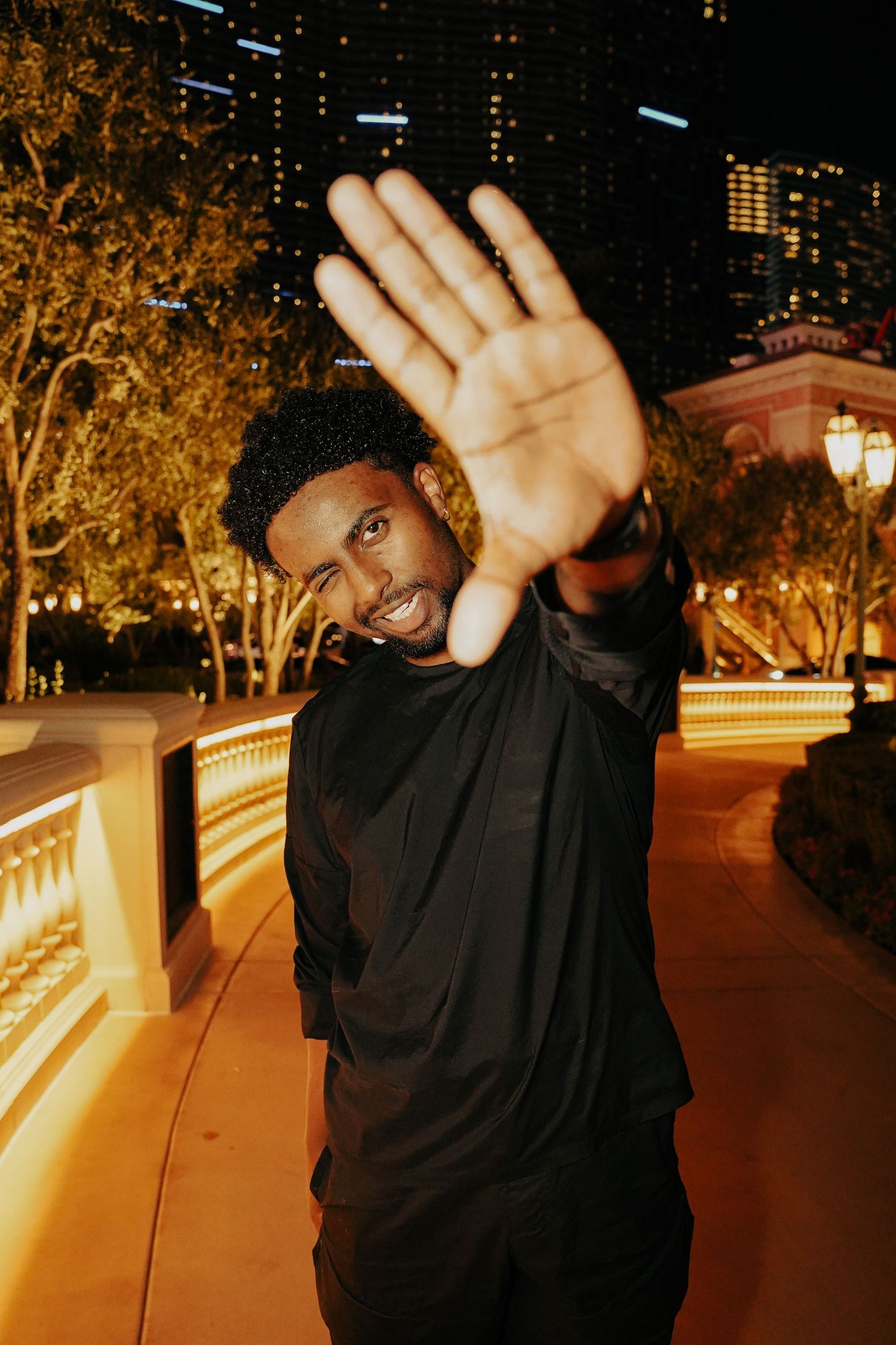 Person in black outfit making stop gesture while smiling at night with lit trees in background.