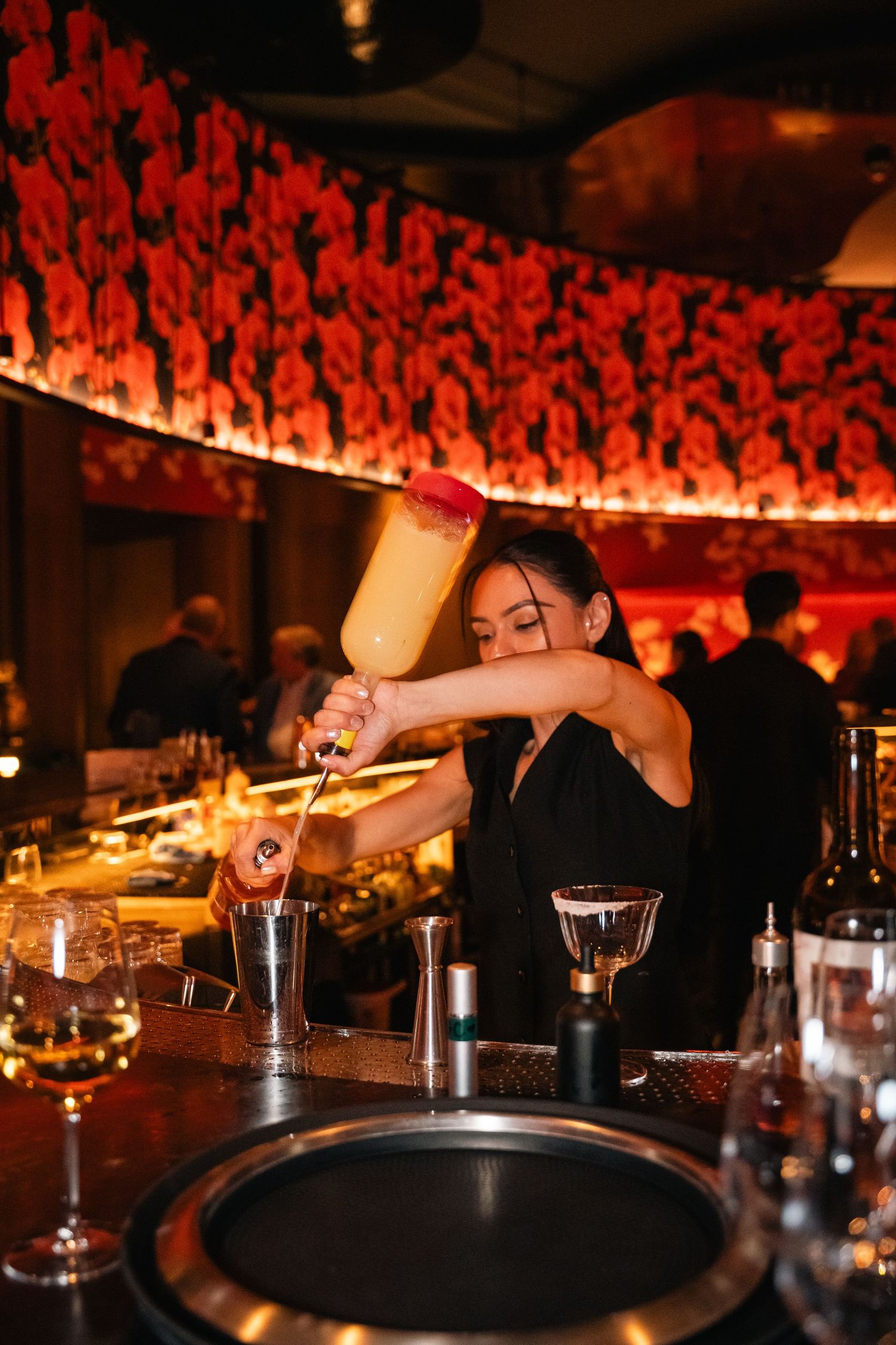 A bartender pouring a cocktail with dramatic red lighting and decor in the background.