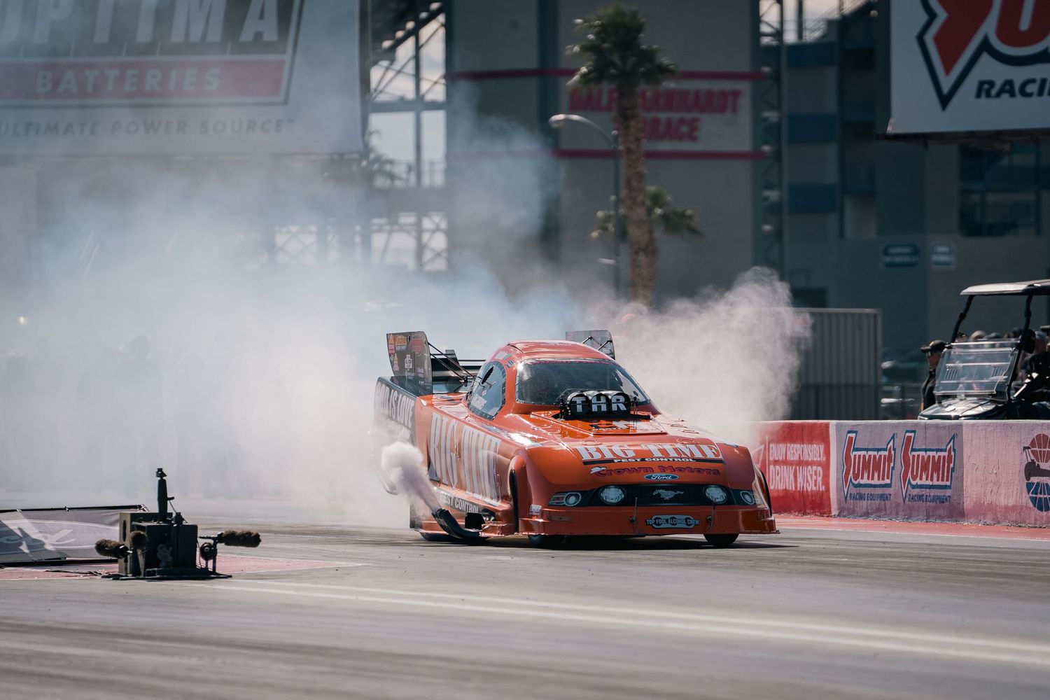 Orange dragster racing car performing a burnout with smoke on a drag strip during competition.
