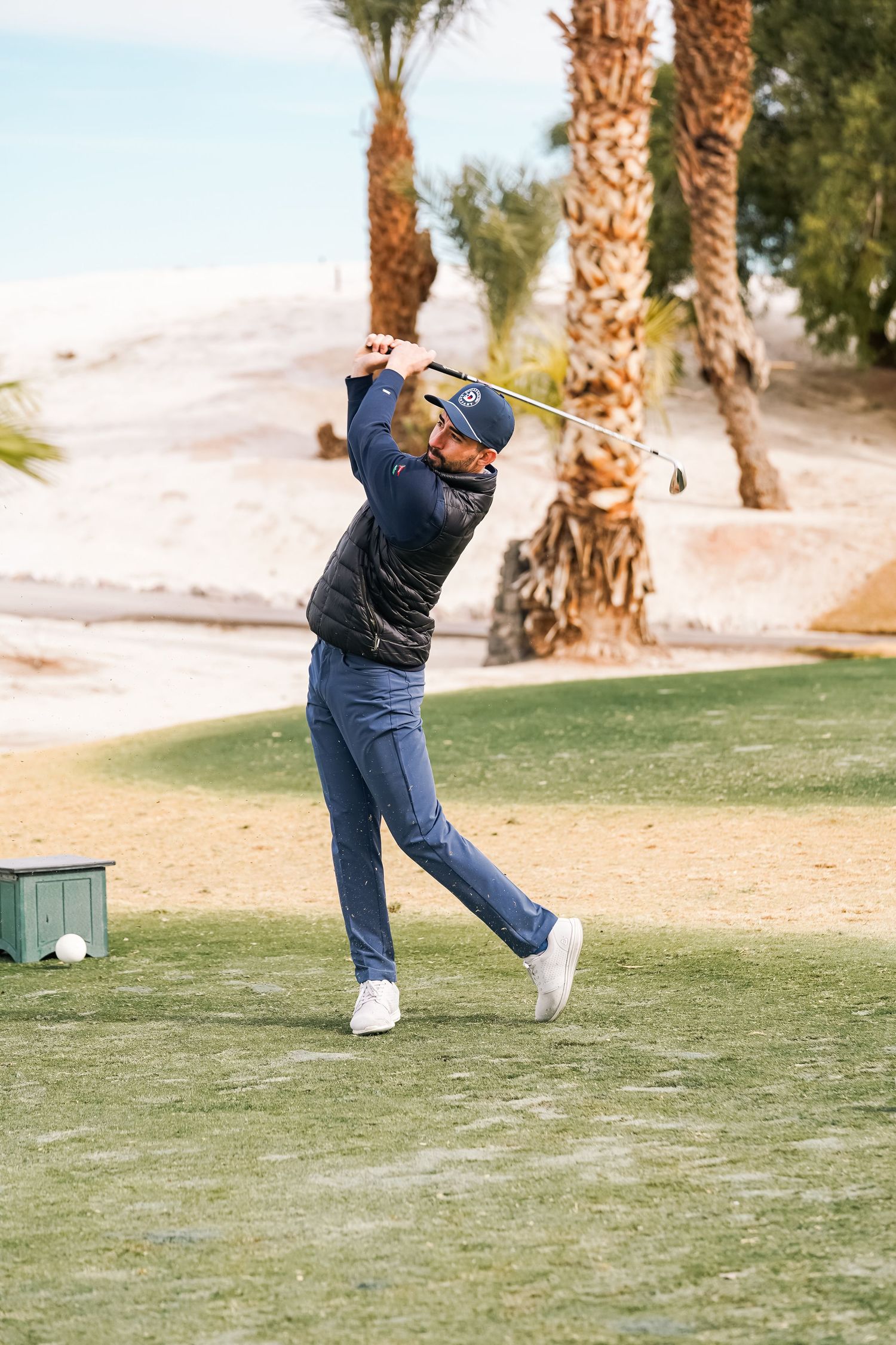 Golfer completes backswing motion on desert golf course with palm trees in background.