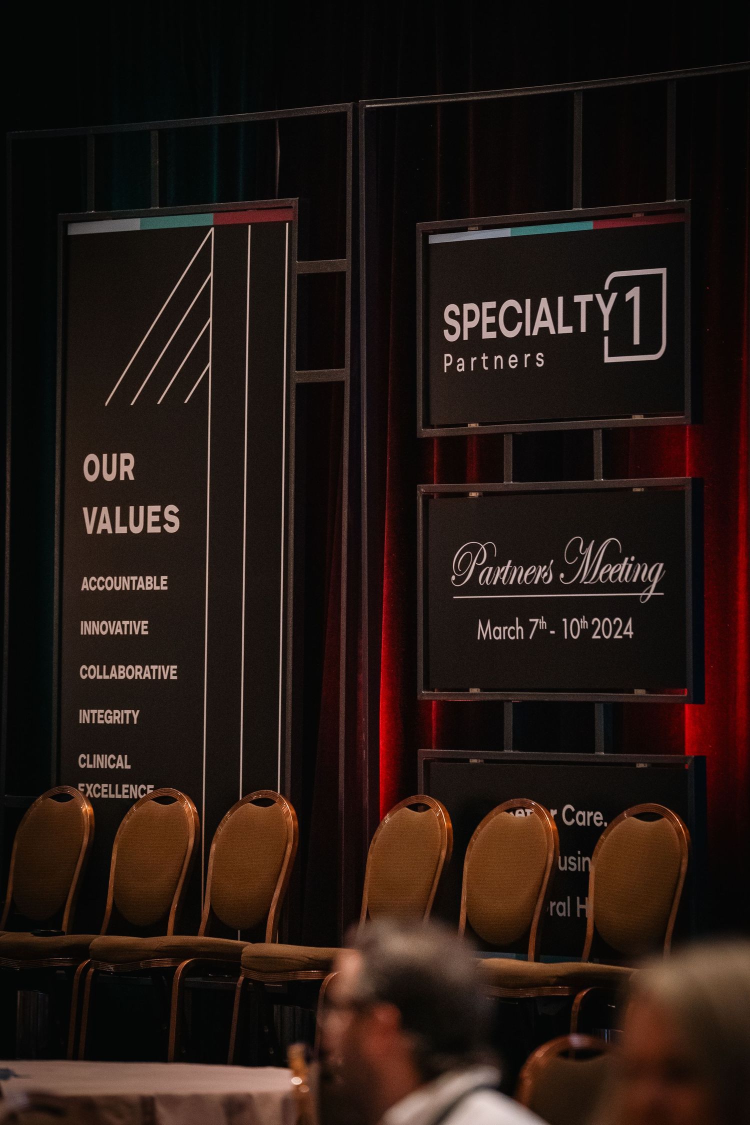 Corporate conference stage displays company values and Specialty1 Partners meeting information.