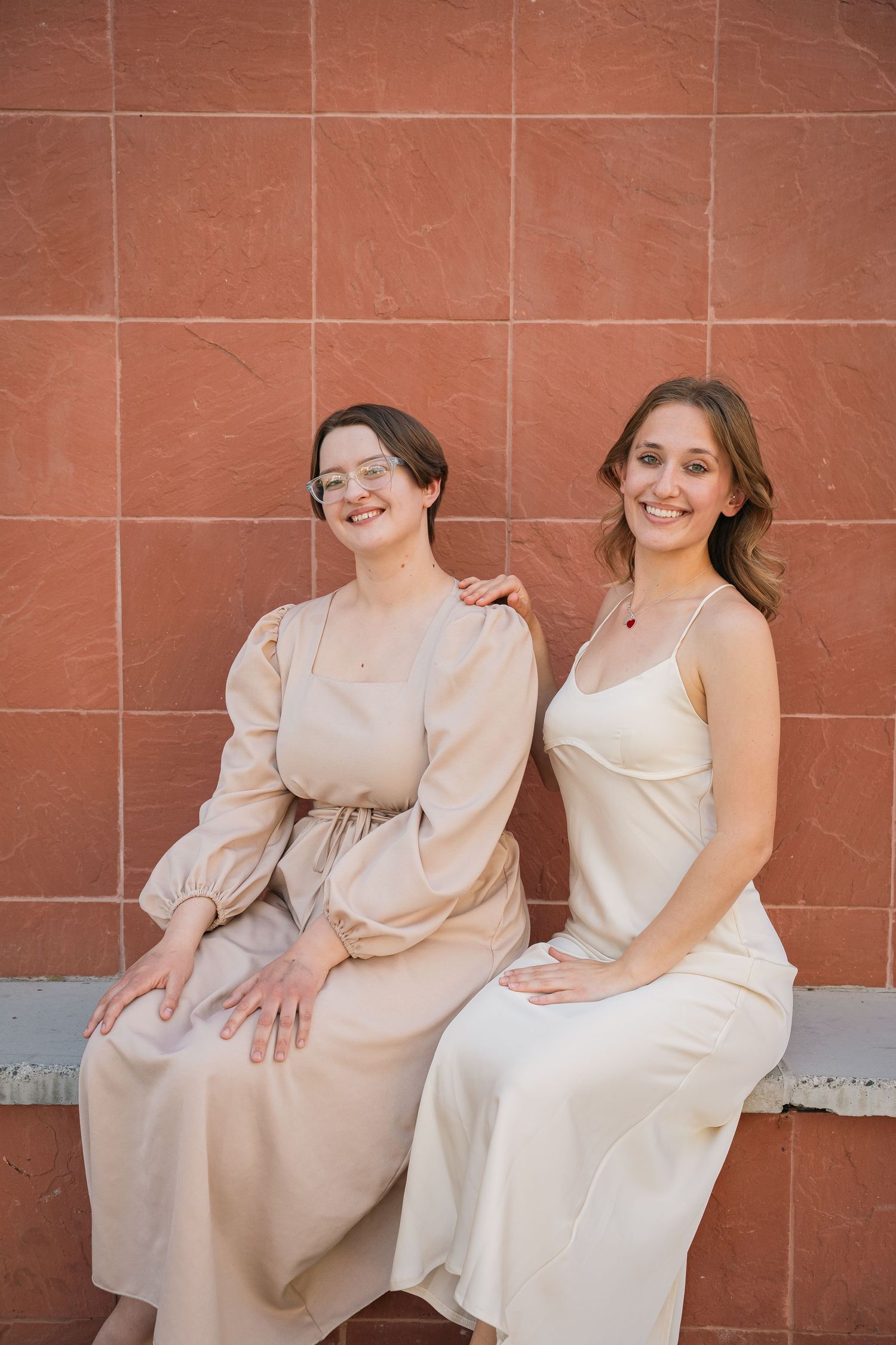 Two friends in neutral-toned maxi dresses sit together on a concrete ledge.
