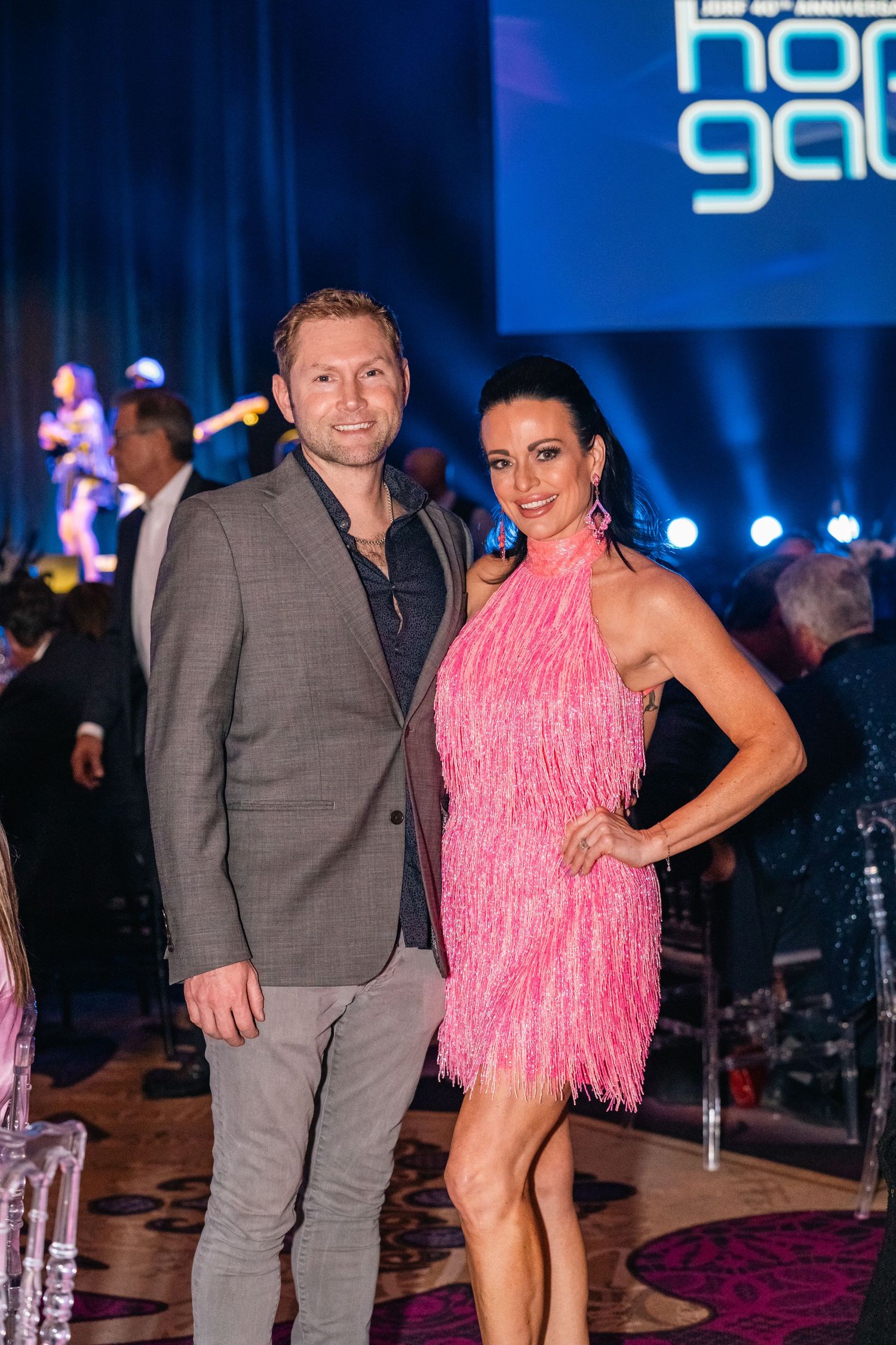 Two attendees at the Hope Gala pose together, with one wearing a grey suit and another in a pink fringed dress.