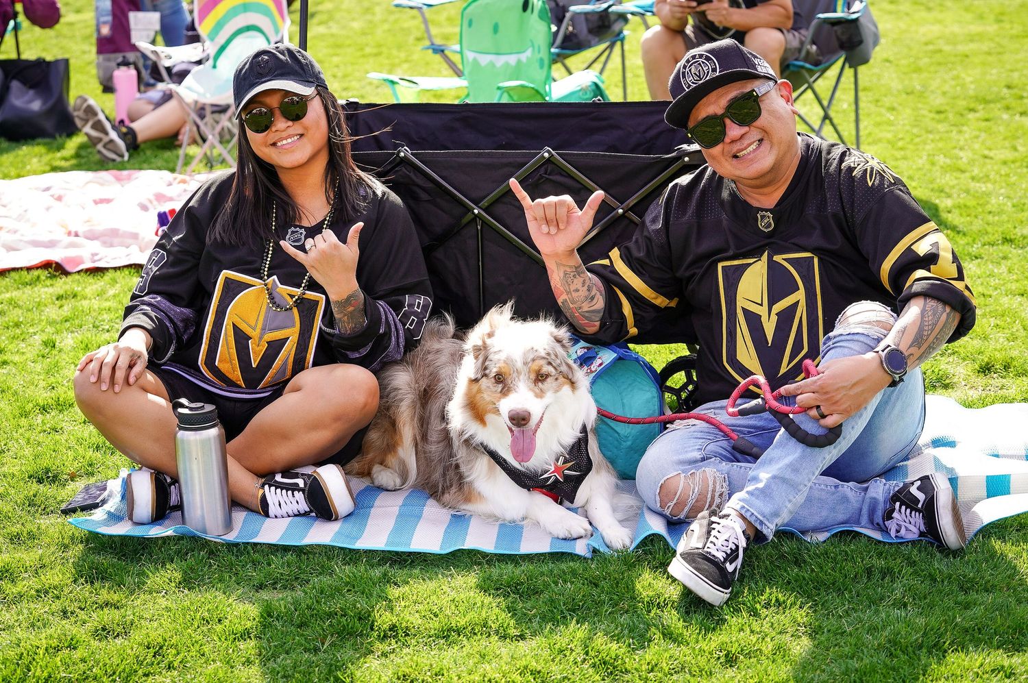 Couple in Vegas Golden Knights jerseys poses with their Australian Shepherd on picnic blanket.