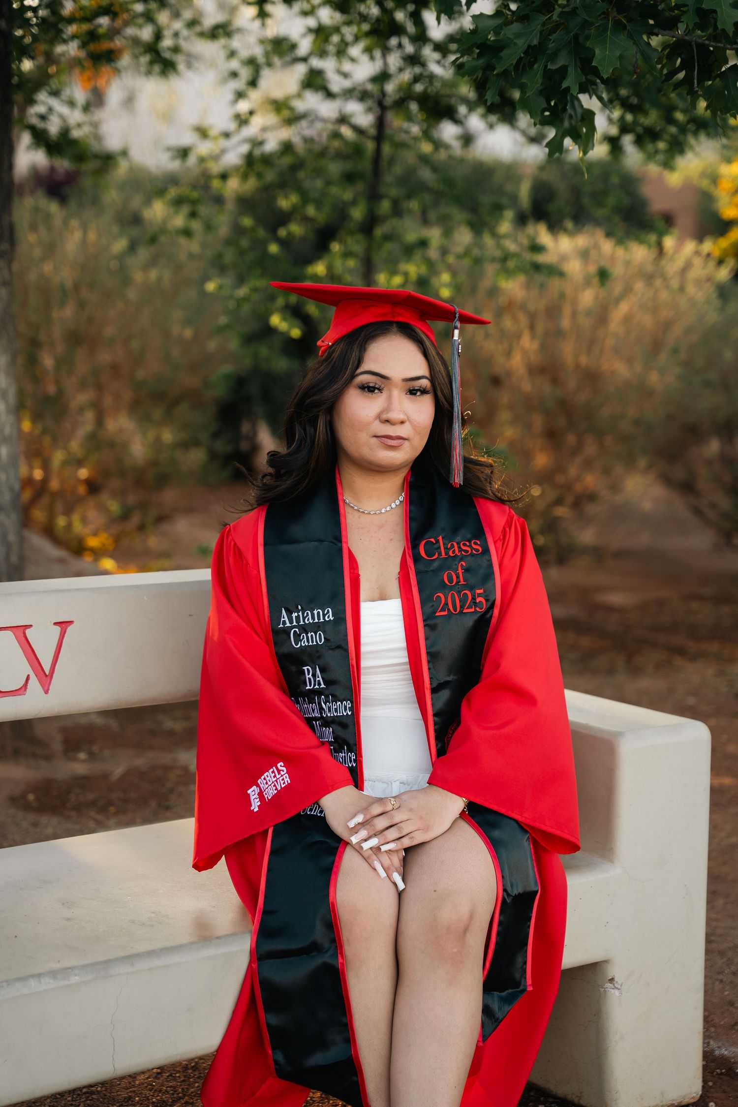 Graduate in red graduation regalia seated on bench during outdoor photo session.