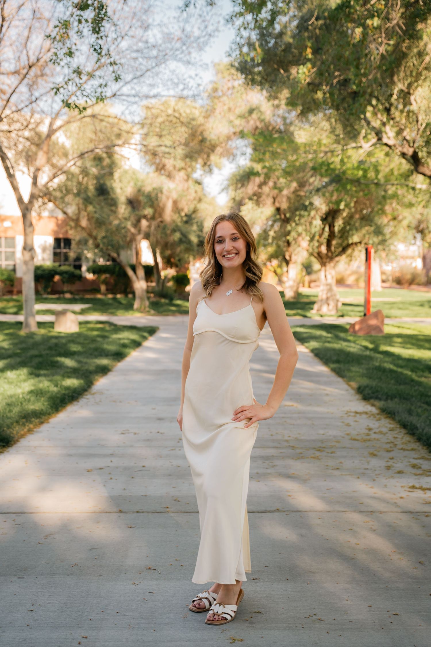 Woman in cream slip dress smiles while posing on tree-lined walkway.