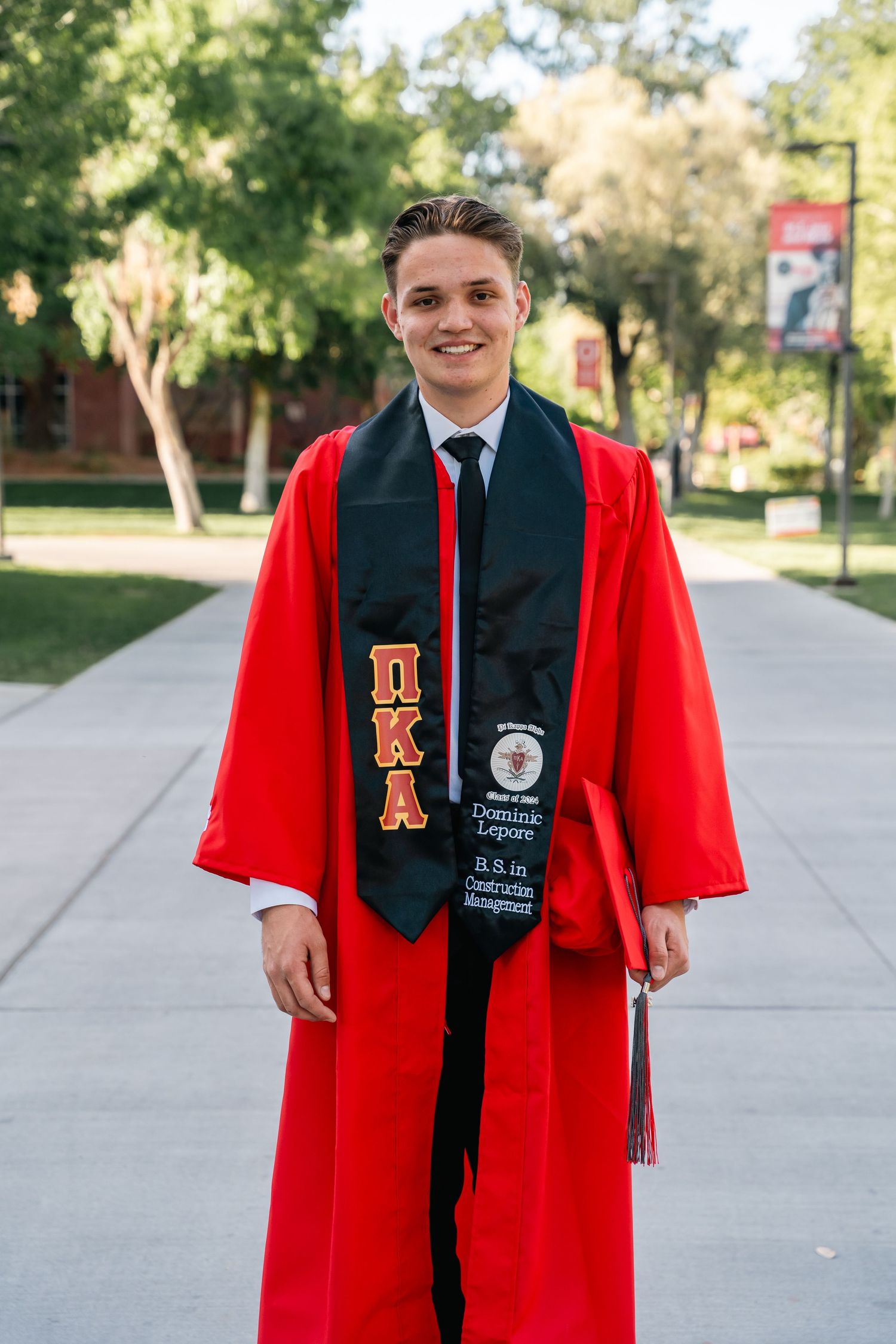 Graduate wearing red graduation gown and decorated sash poses on campus pathway.