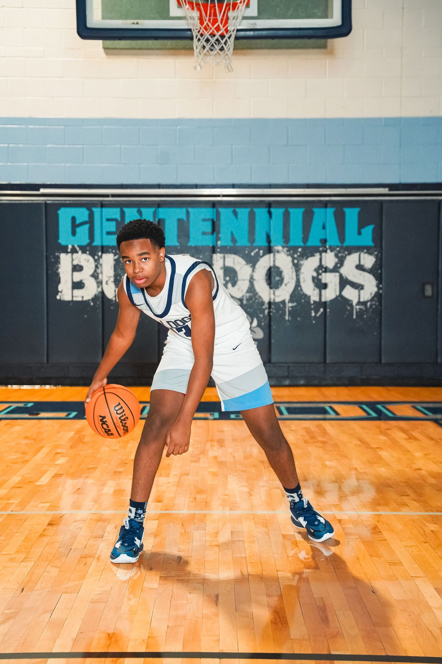Player demonstrates low dribbling stance on basketball court with Centennial Bulldogs branding.