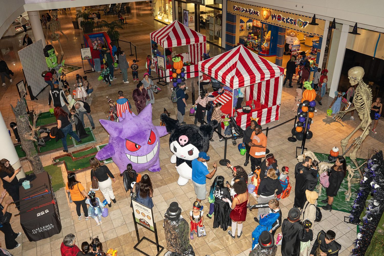 An aerial view of a mall Halloween event with striped tents, purple lighting, and crowds of costumed attendees.