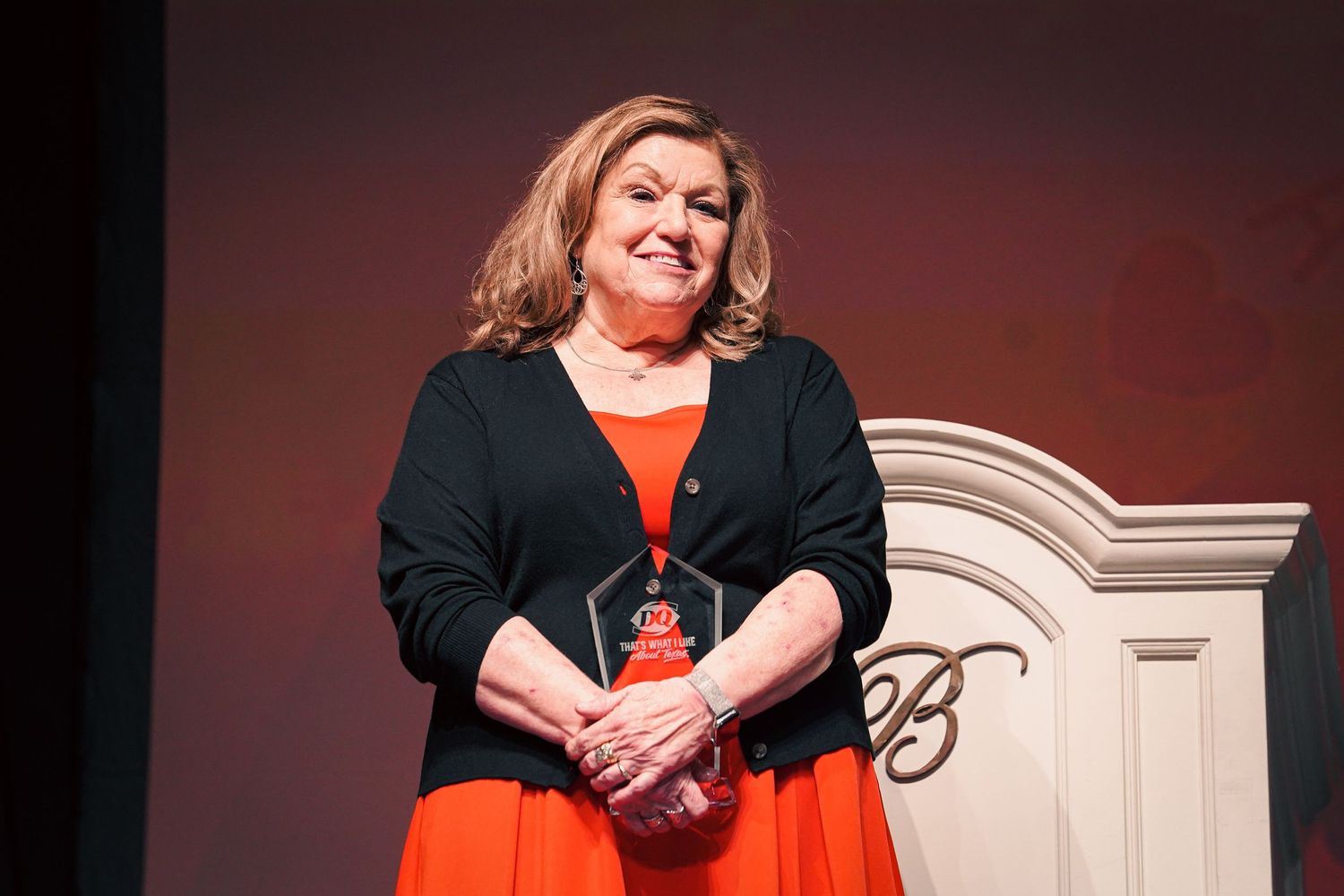 Woman in black cardigan and orange dress smiles on stage at Vegas conference.