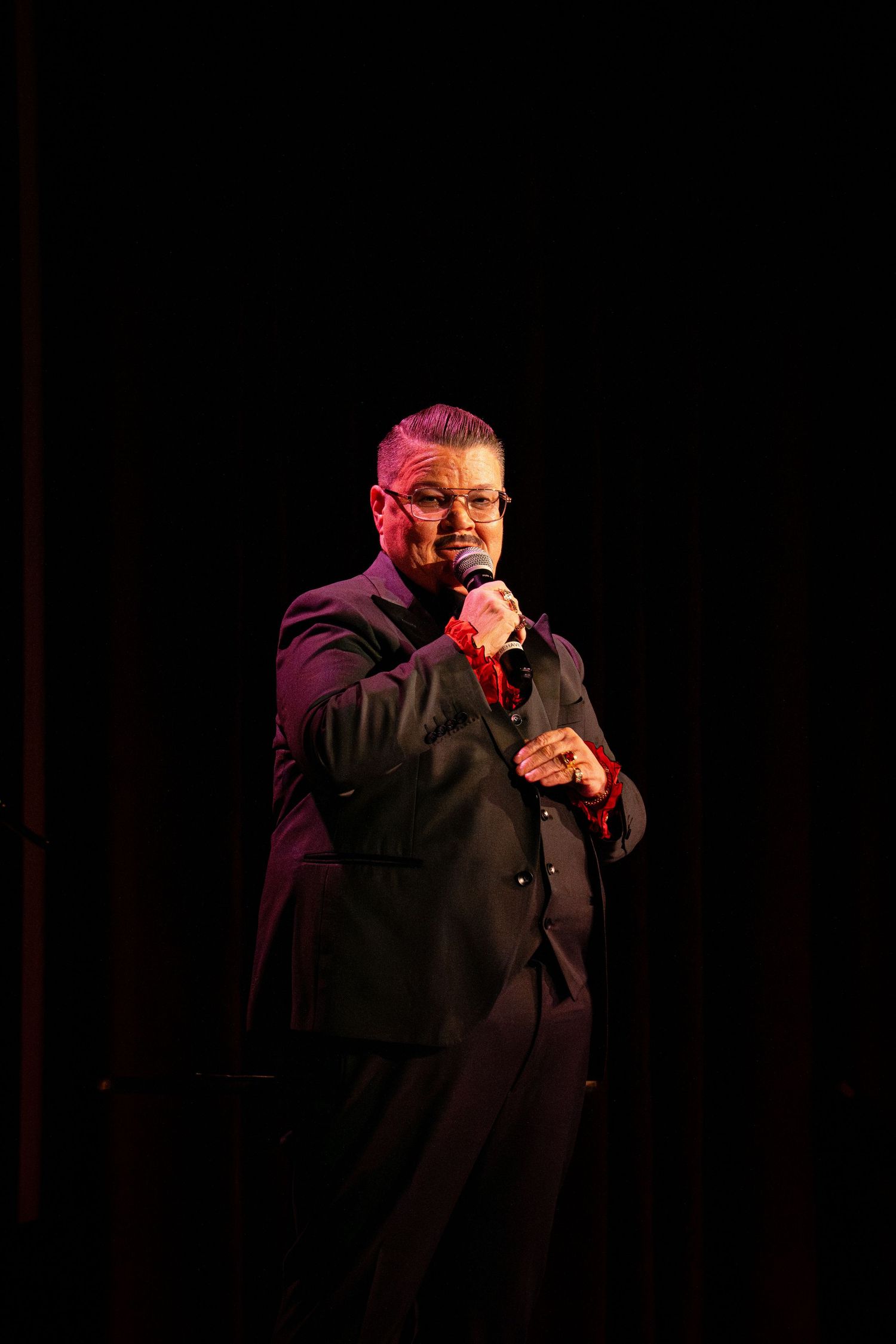 A performer in a black suit holds a microphone while performing on a darkened stage.