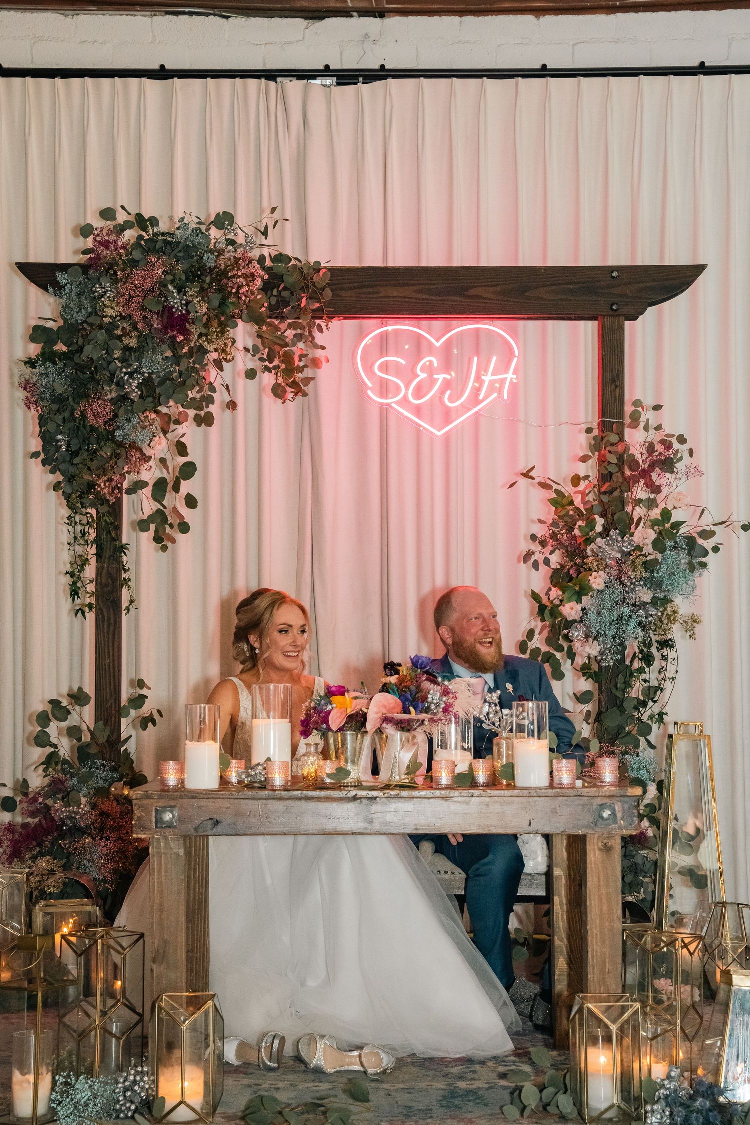 Wedding couple sits at sweetheart table decorated with floral arch and neon heart sign against white backdrop.
