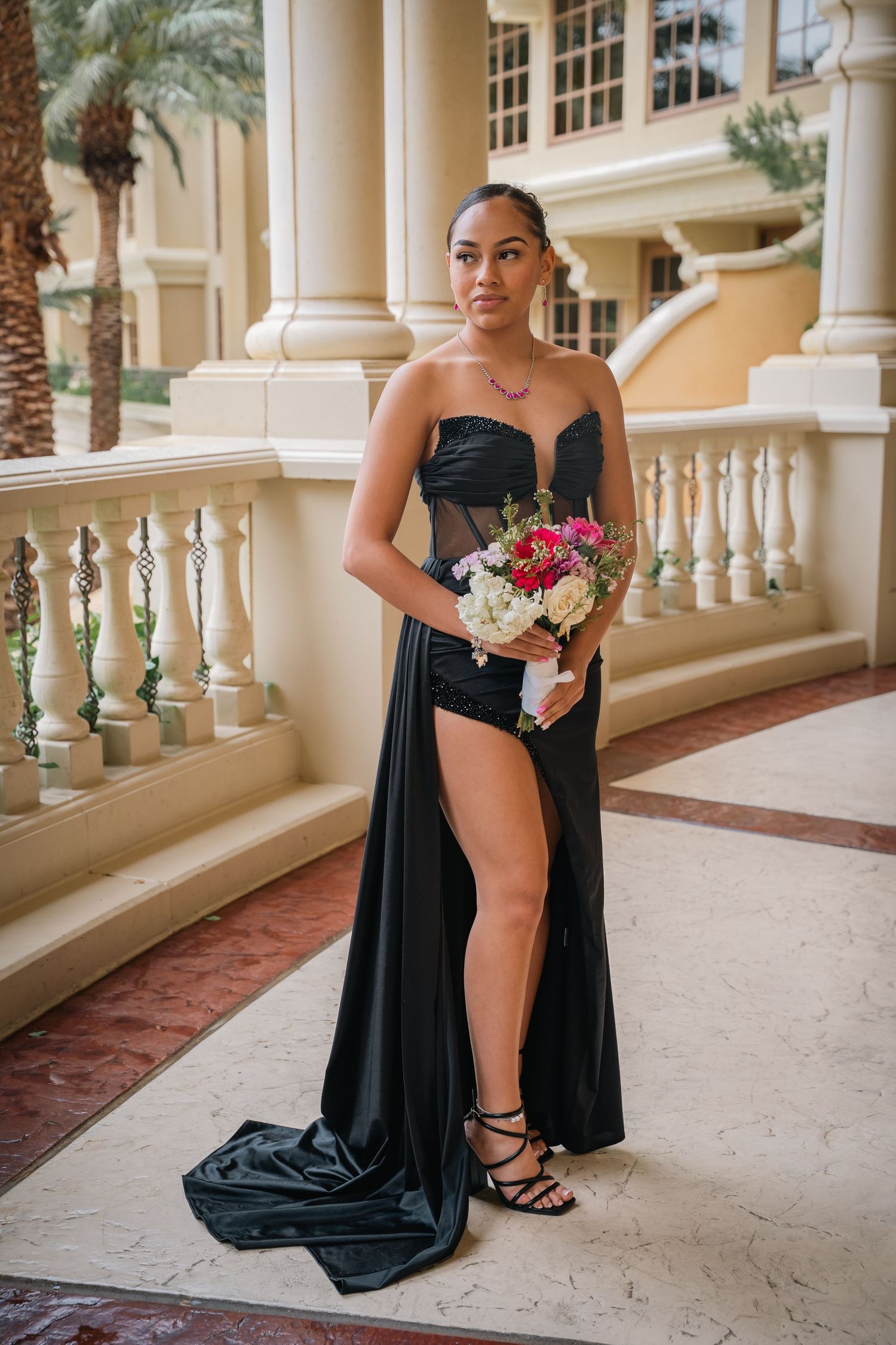A person in a strapless black gown holds a bouquet while posing by elegant columns.