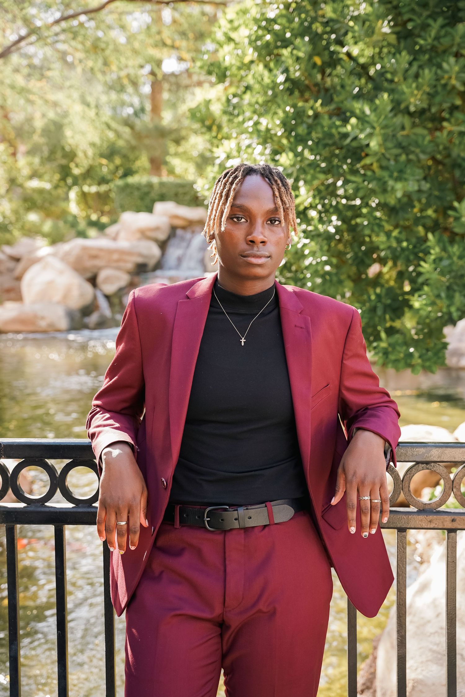 A person wearing a burgundy suit and black turtleneck poses on an outdoor stone balcony near a waterfall.