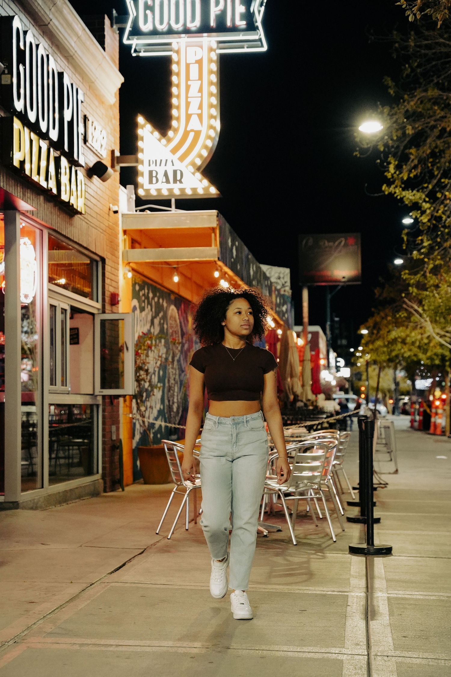 Woman strolling past illuminated Good Pie restaurant sign on city street at night.