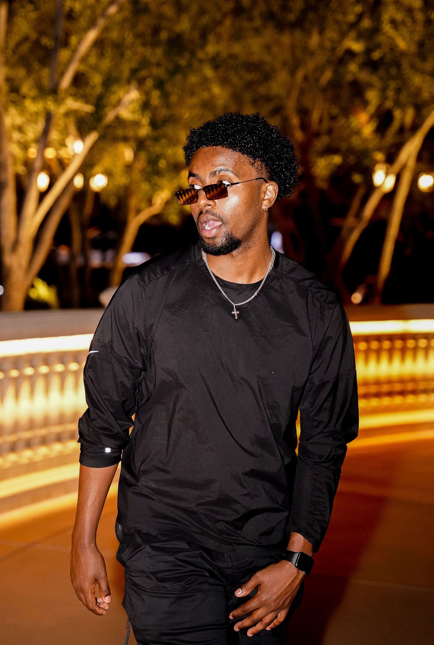 Person in black attire and sunglasses posing outdoors at night with illuminated trees.