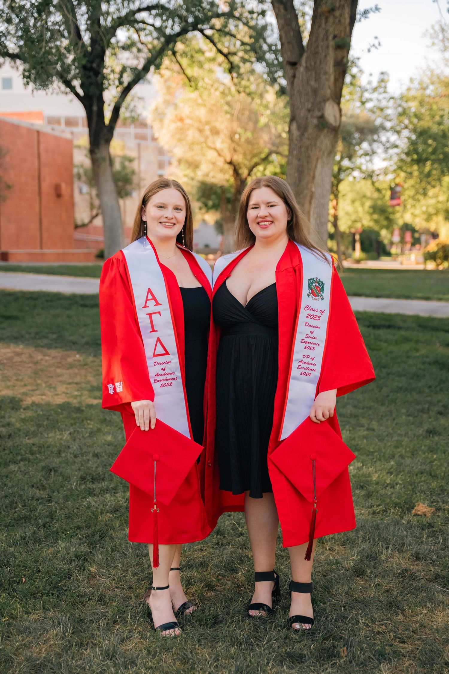 Two graduates in red graduation robes and sashes pose together on campus lawn.