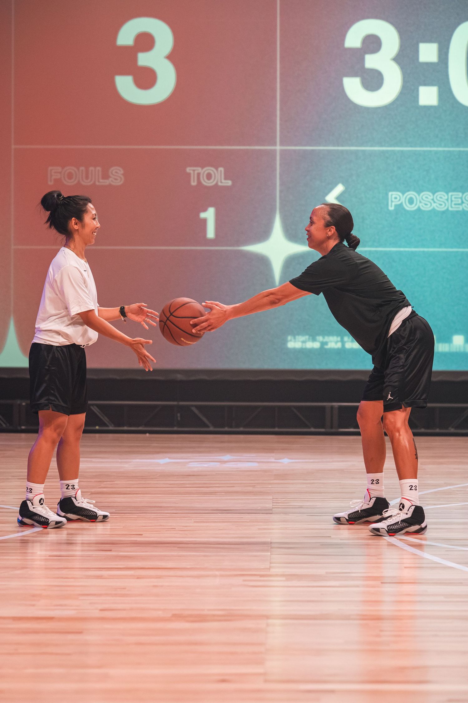 Two basketball players pass a ball during practice on an indoor court with a scoreboard display.