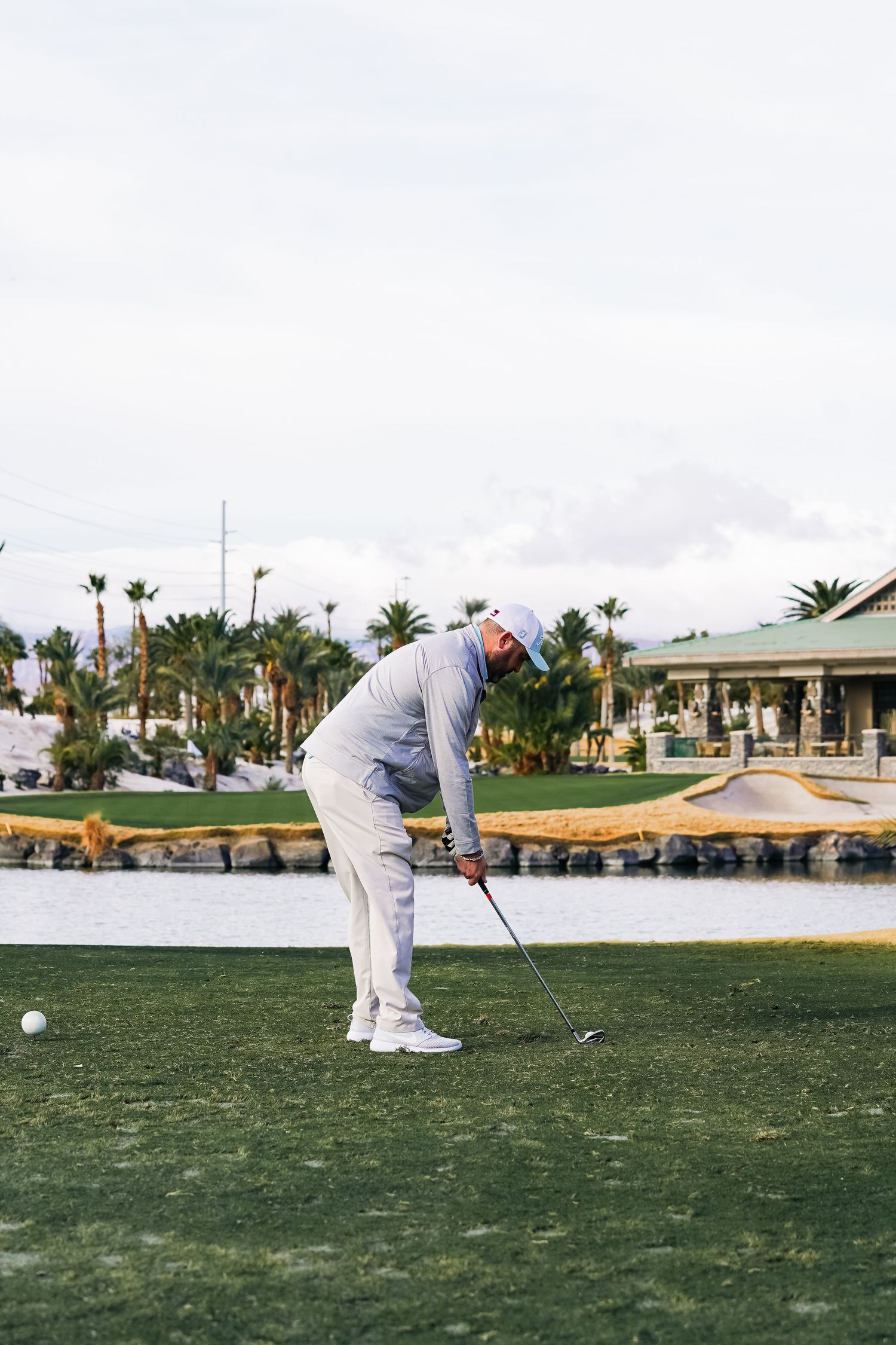 Golfer lines up a putt on lush green course with water hazard and palm trees in background.