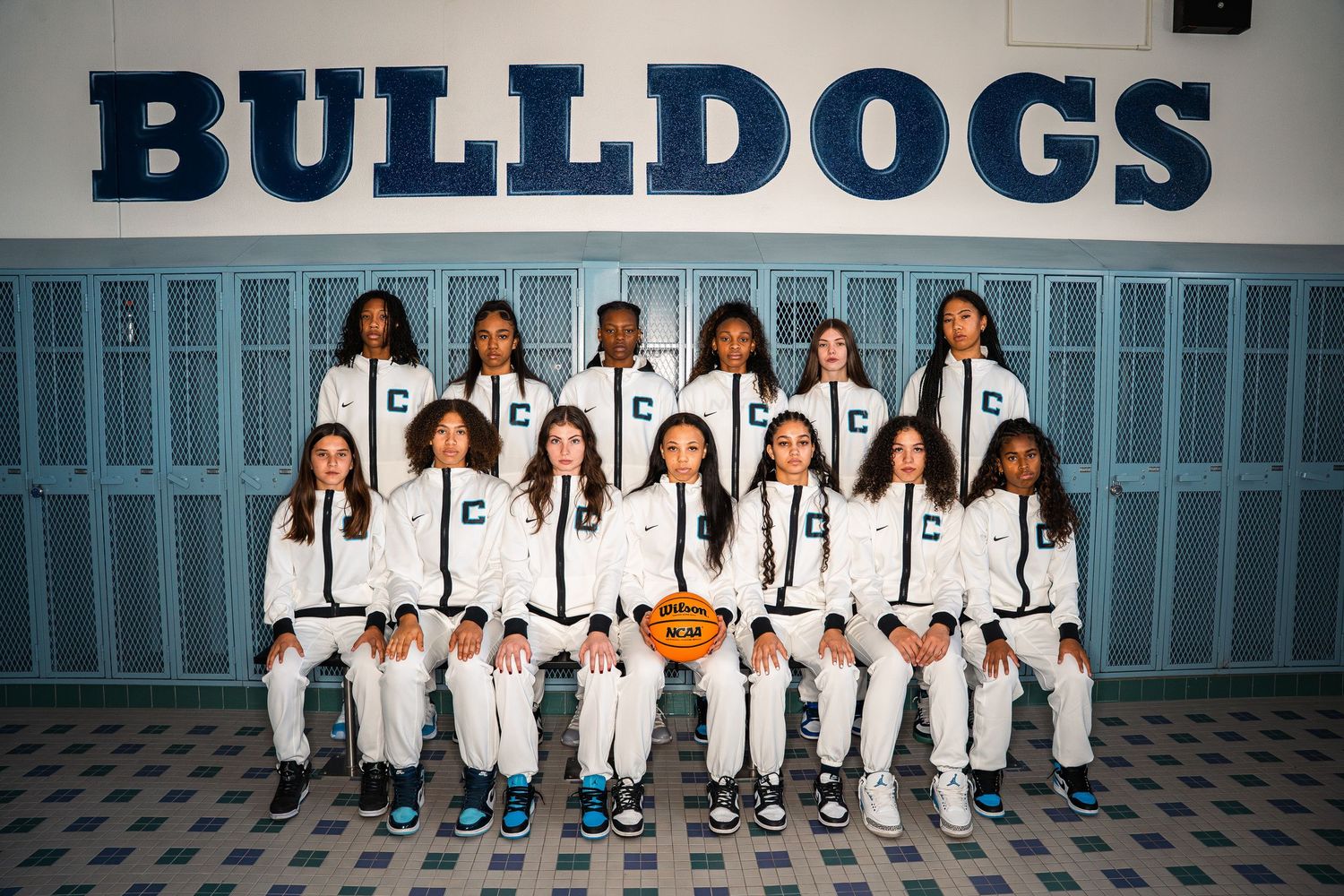 Team photo of basketball players in white warm-up uniforms against school wall.