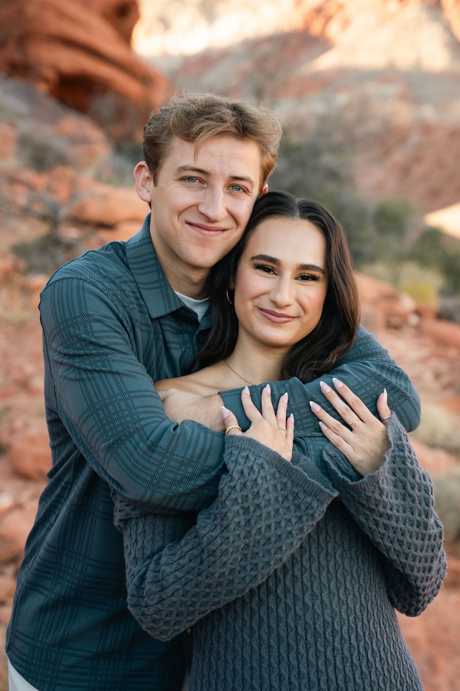 Couple embracing and showing engagement rings against red rock backdrop.