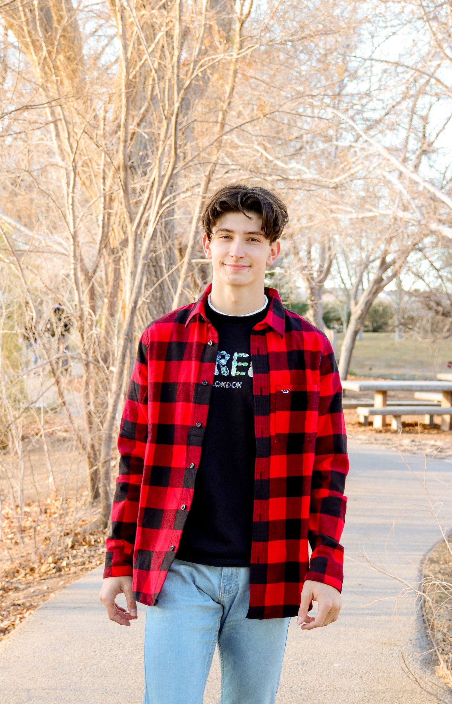 Young person in red plaid flannel and jeans stands on pathway with bare trees in background.