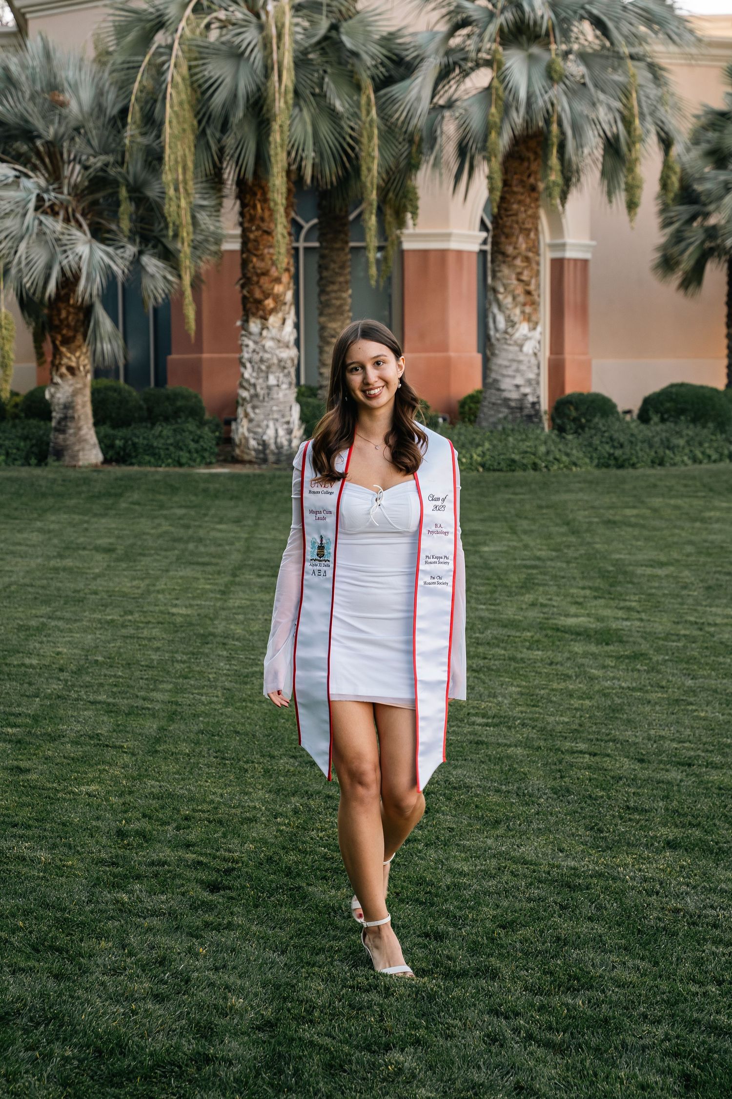 White dressed person poses on grass lawn with tall palm trees and Mediterranean style buildings in background.