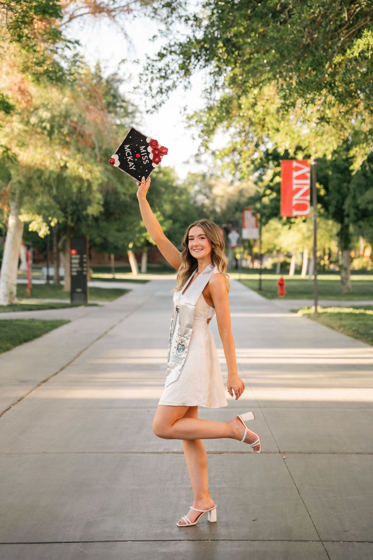 Graduate celebrates on campus walkway surrounded by trees in outdoor graduation photoshoot.