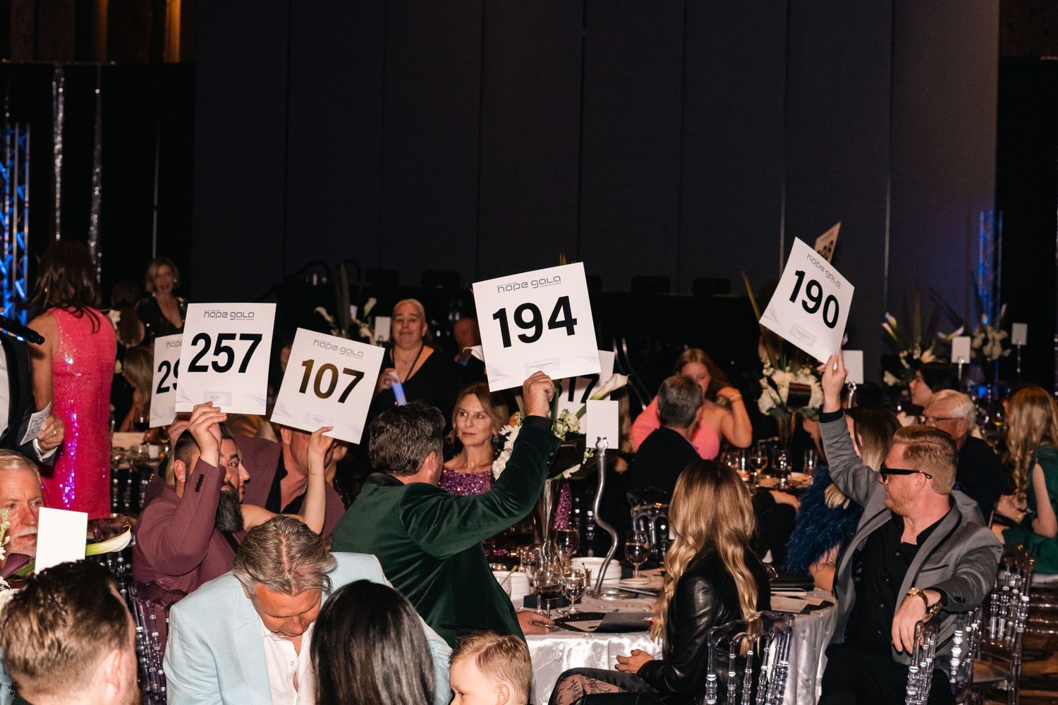 Crowd holding up numbered bid cards during an auction event in a dimly lit venue.