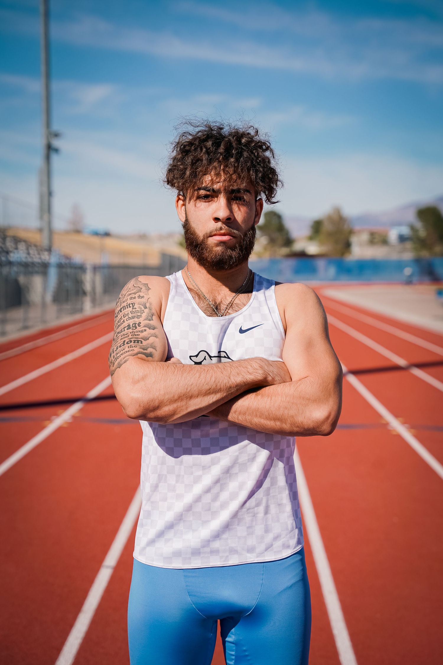 Athlete in white Nike tank and blue shorts stands with crossed arms on track field.