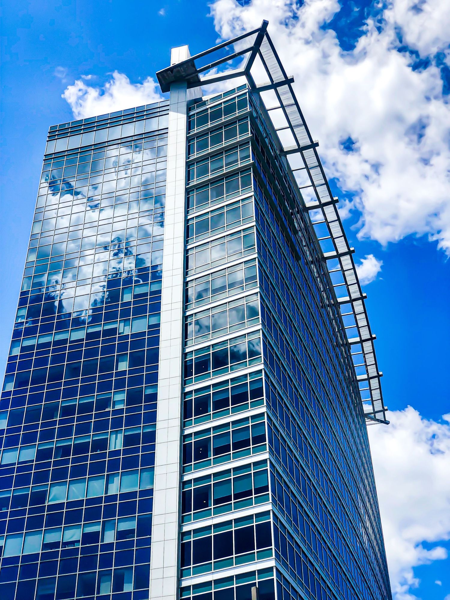 Modern glass skyscraper reflecting clouds in blue sky with geometric architectural details.