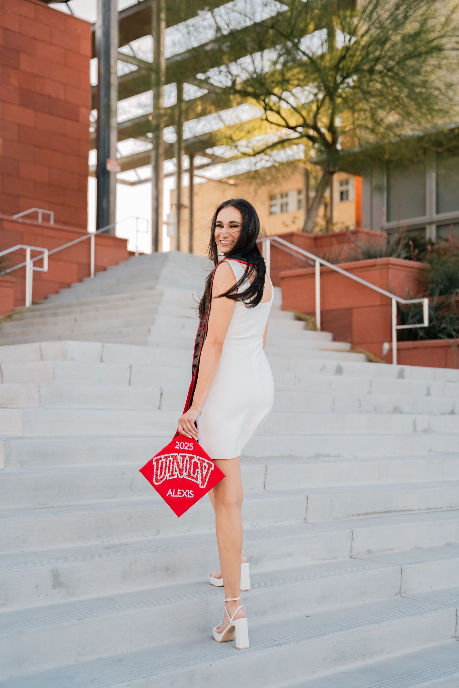 Graduate in white dress poses with decorated graduation cap on campus steps.