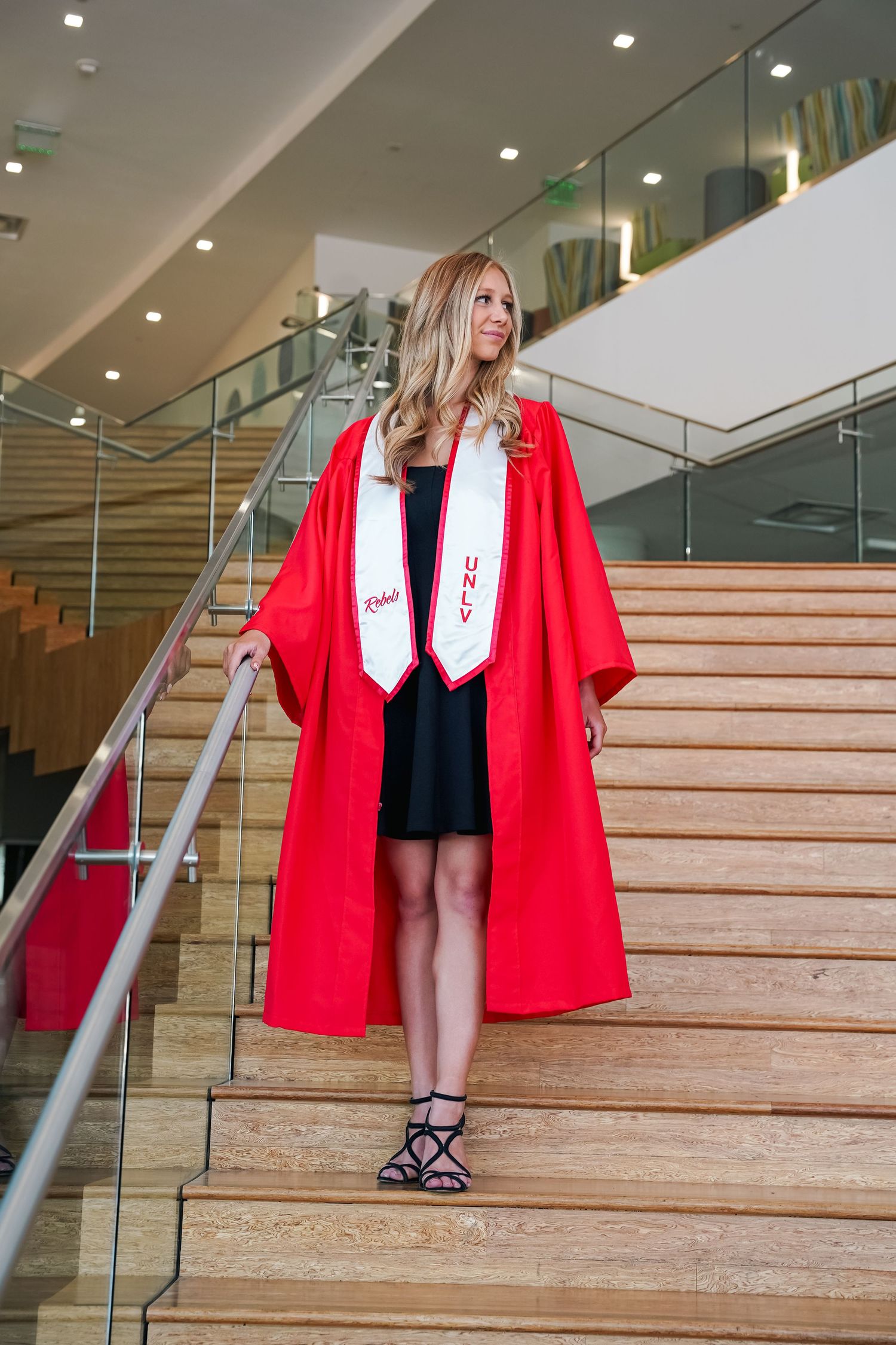 Graduate wearing red graduation gown stands proudly on wooden stairs with glass railing.