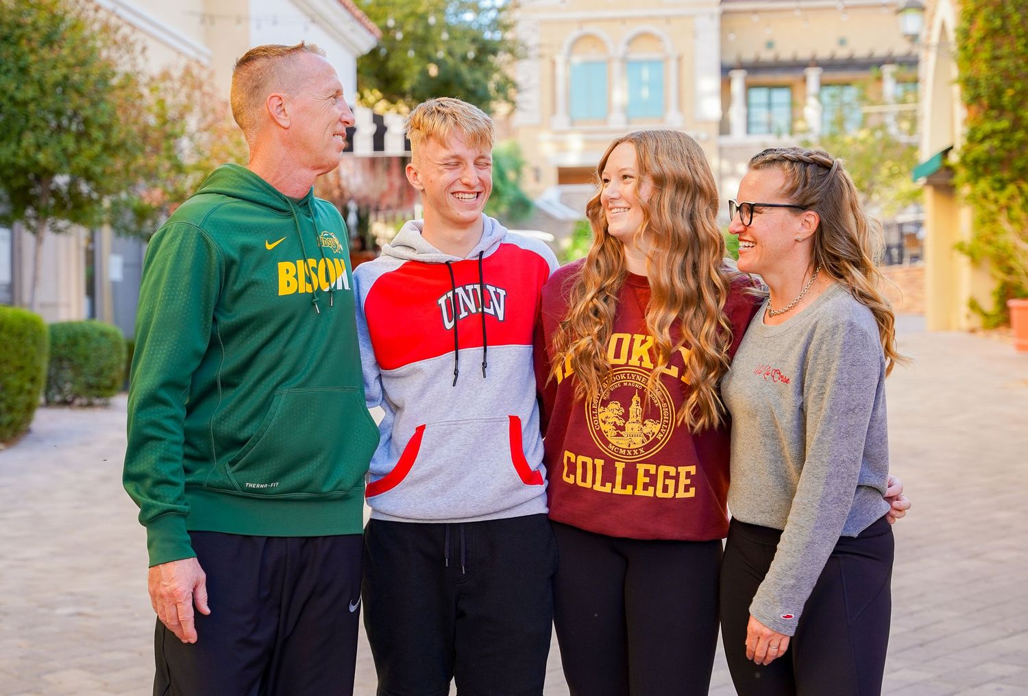 Four people wearing college sweatshirts pose together on a sidewalk in front of a historic building.
