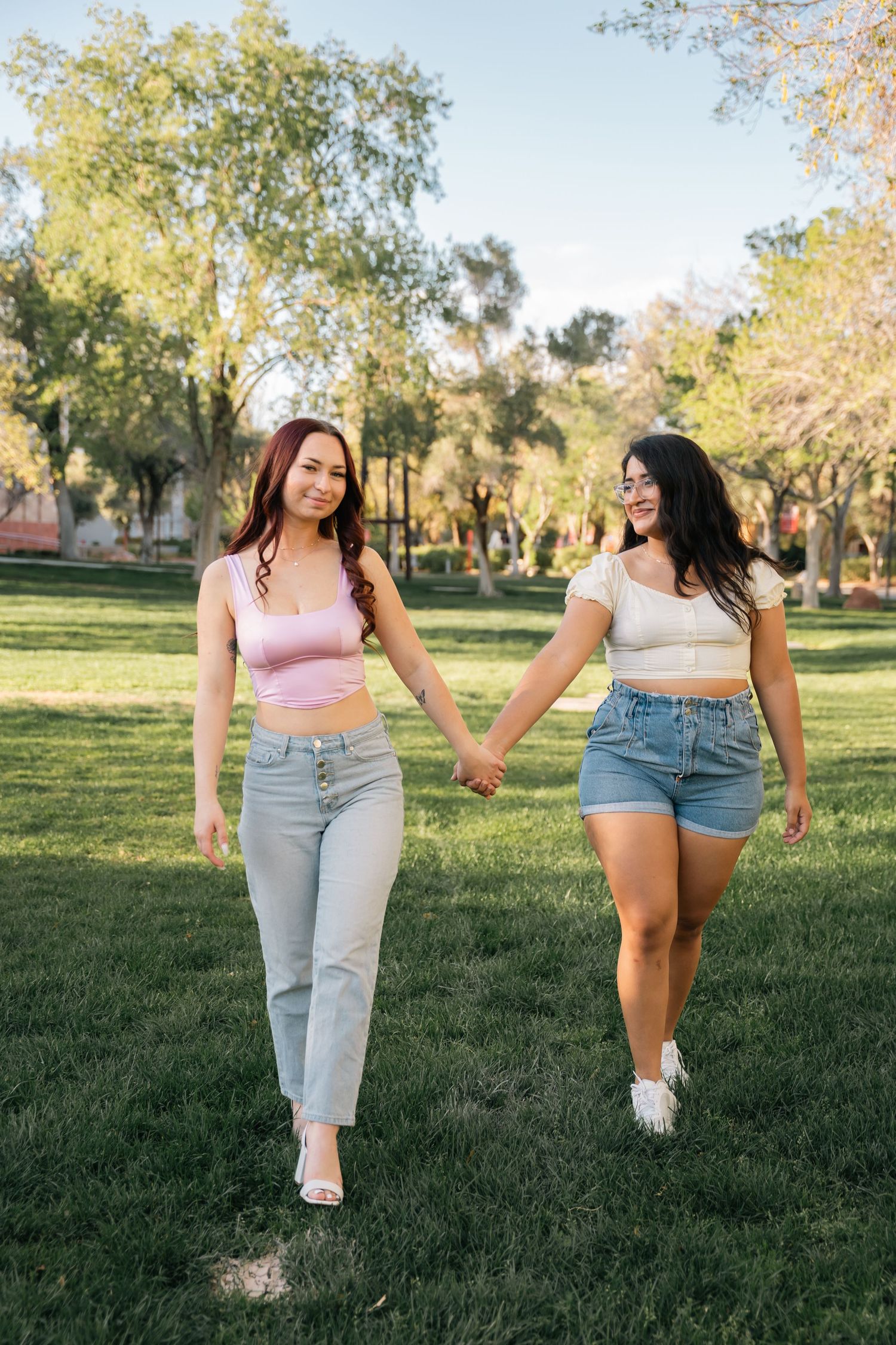 Two friends hold hands while walking together on a grass lawn wearing crop tops and denim.