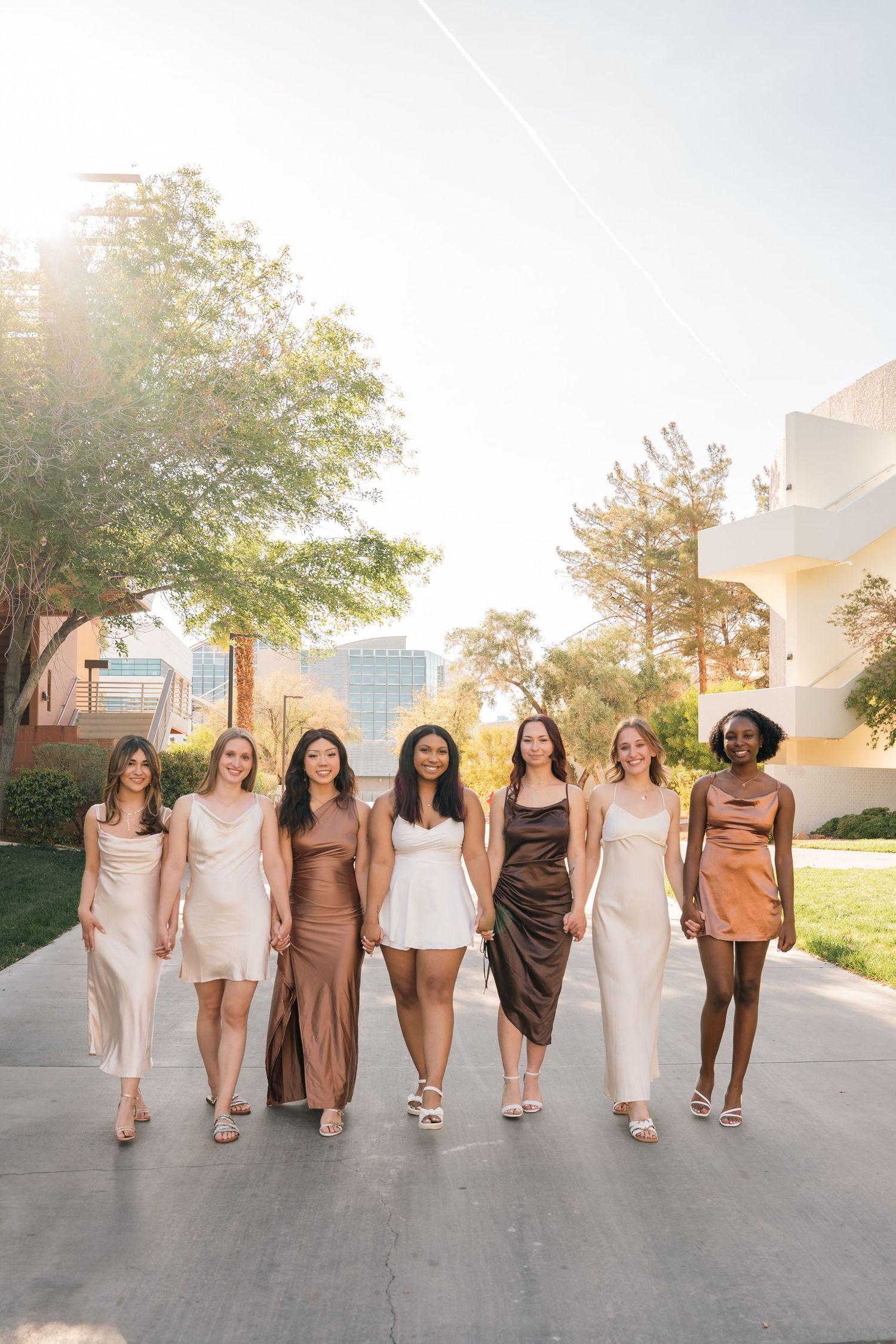 Seven women in coordinated neutral-toned dresses walk together on a sunny pathway.