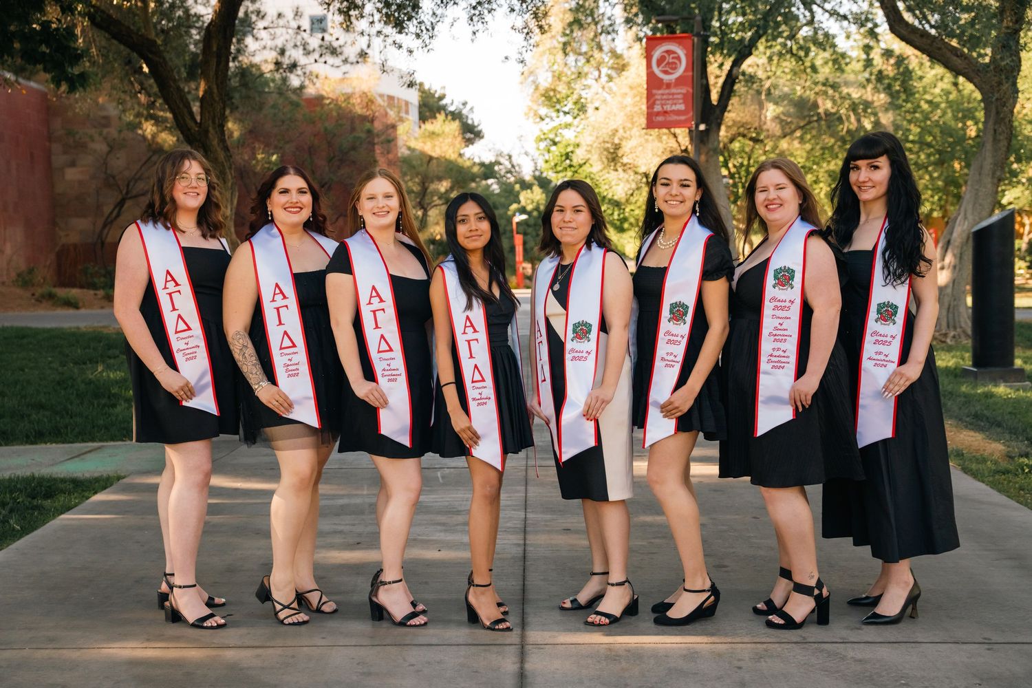 A formal group photo shows people wearing black dresses and graduation sashes standing in a line.
